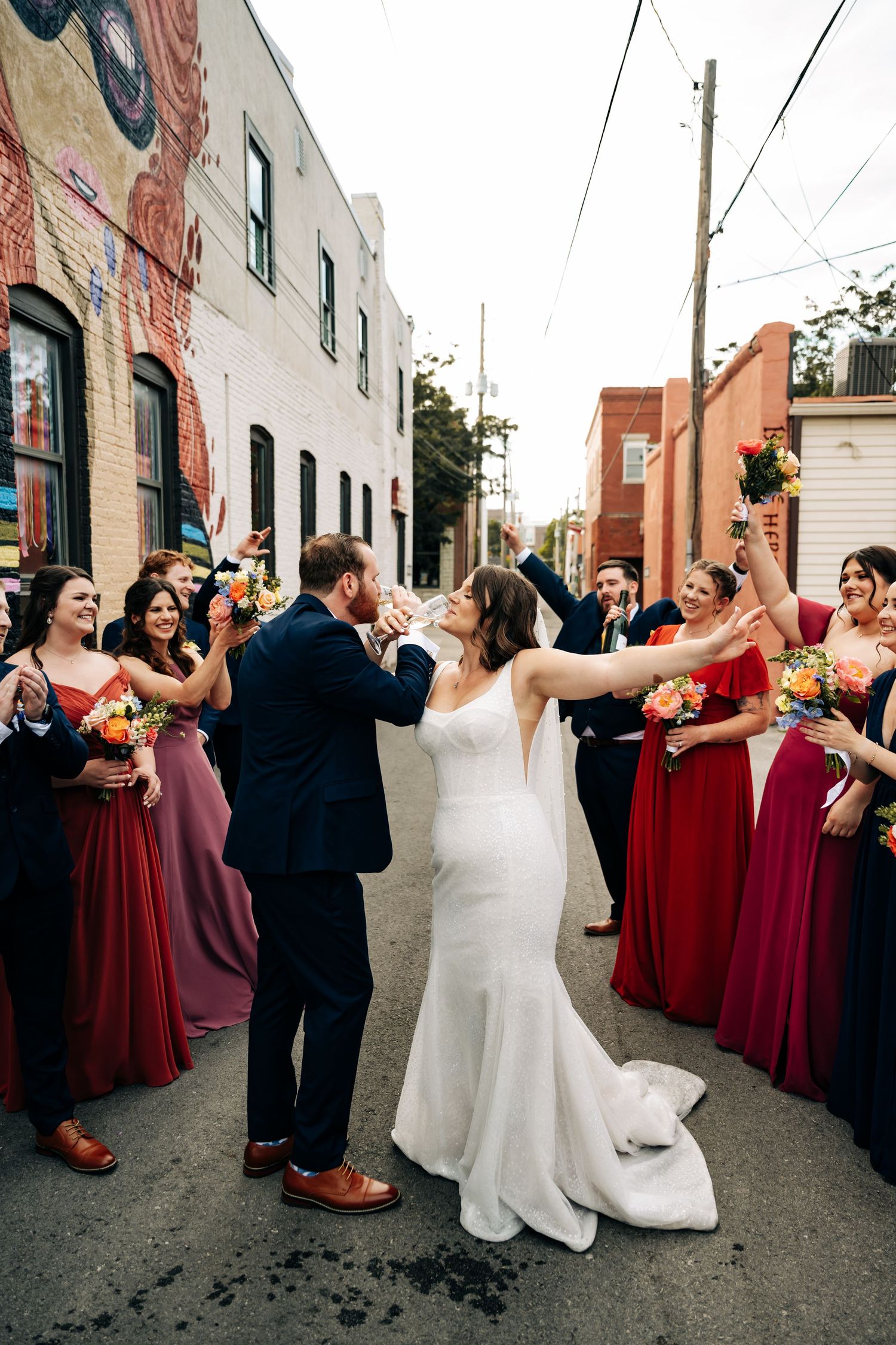 Wedding party lines up in burgundy and black attire for celebratory photos in urban alley.