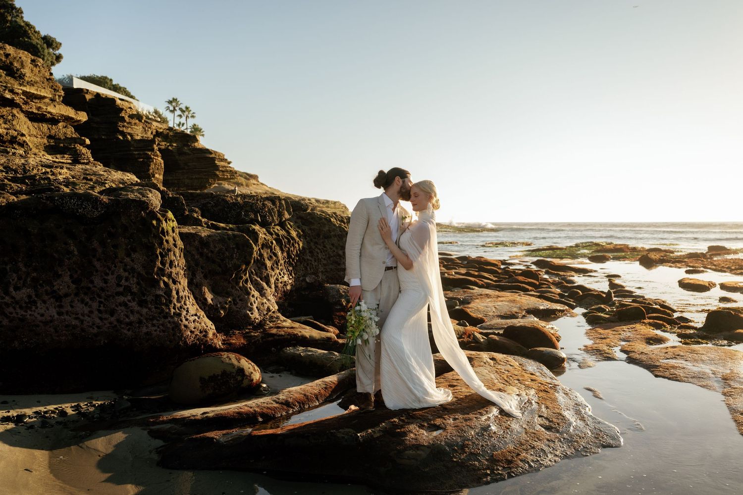 Romantic coastal wedding photoshoot with dramatic rocky cliffs and ocean waves at sunset.