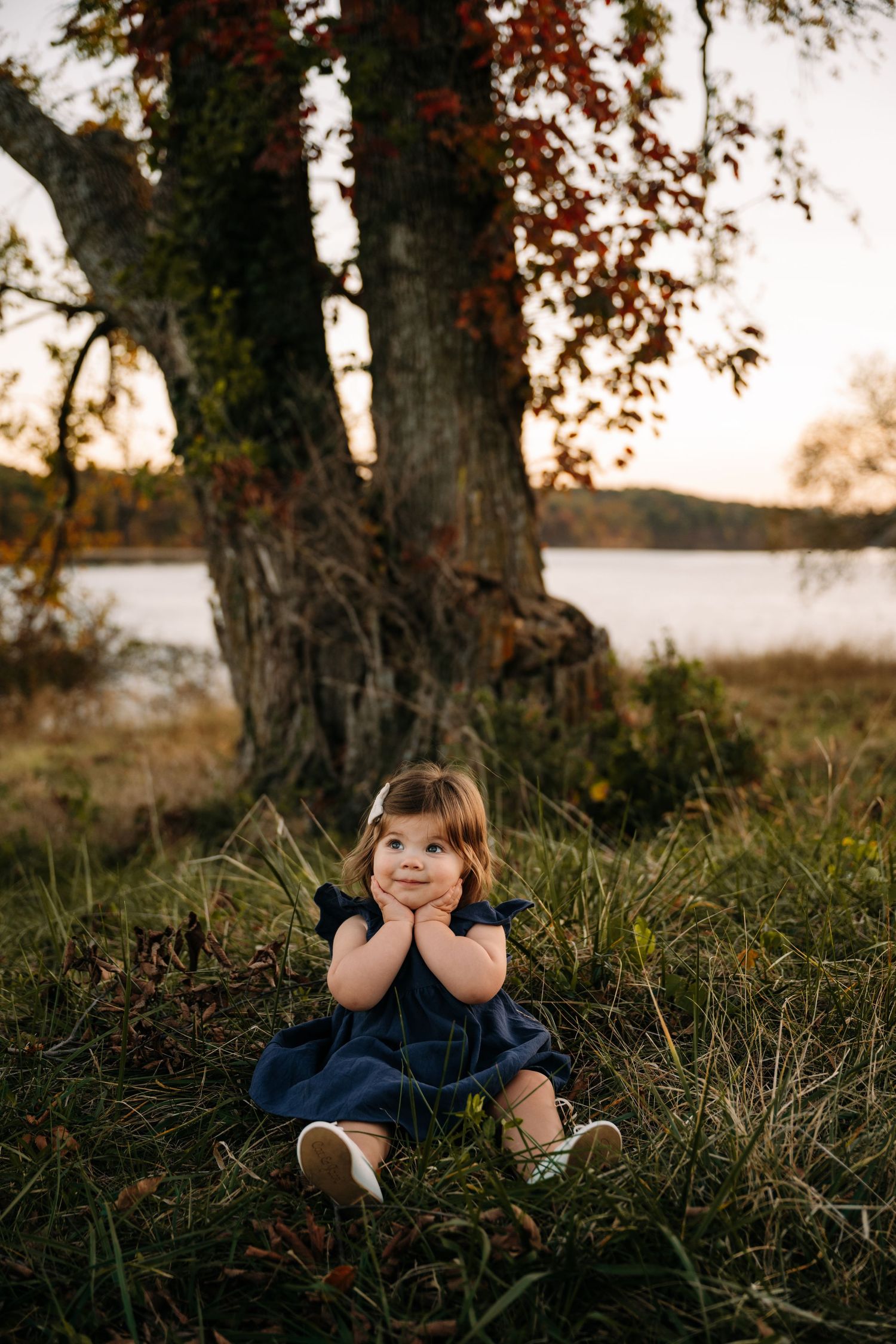 Child sitting in tall grass at sunset near trees in outdoor portrait setting.