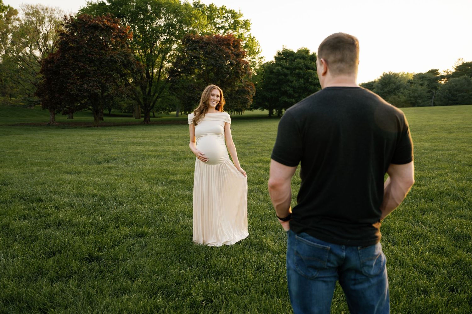 Pregnant woman in white dress poses in green field at golden hour while man in black shirt watches.