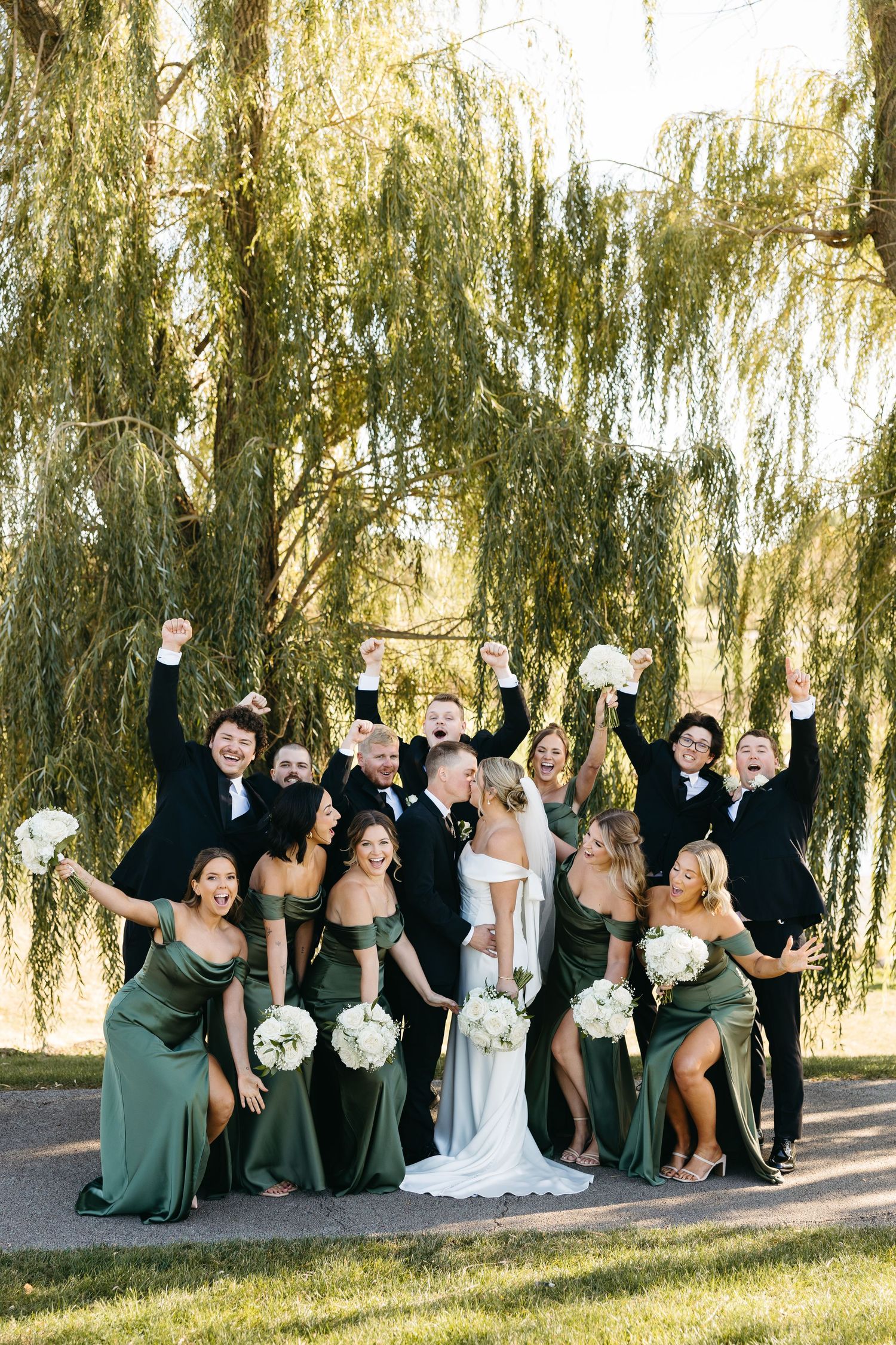 Wedding party in green dresses and black suits poses playfully on grass near weeping willow trees.