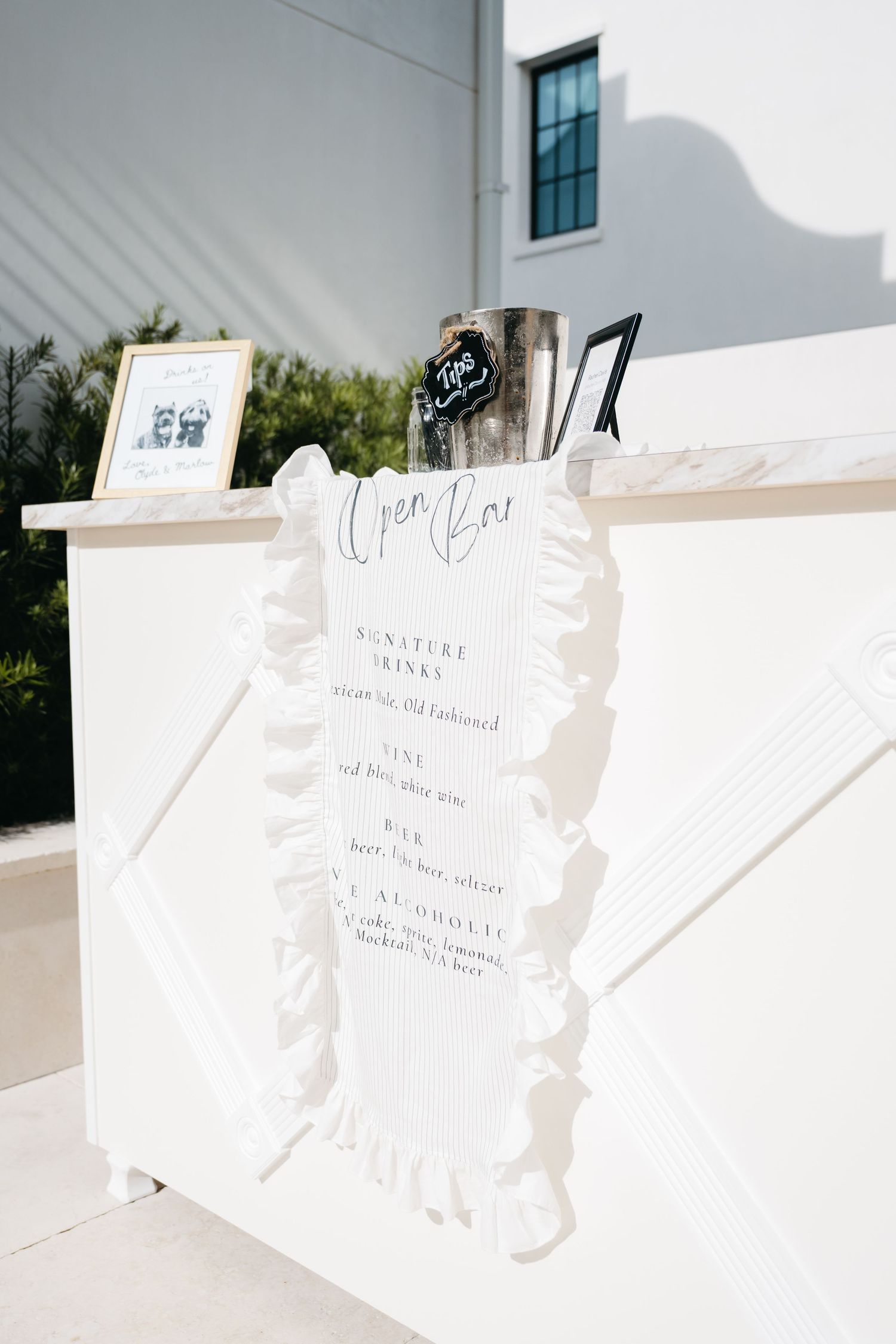 Elegant outdoor open bar display with white ruffled sign listing signature drinks, beer, and non-alcoholic options at a wedding.