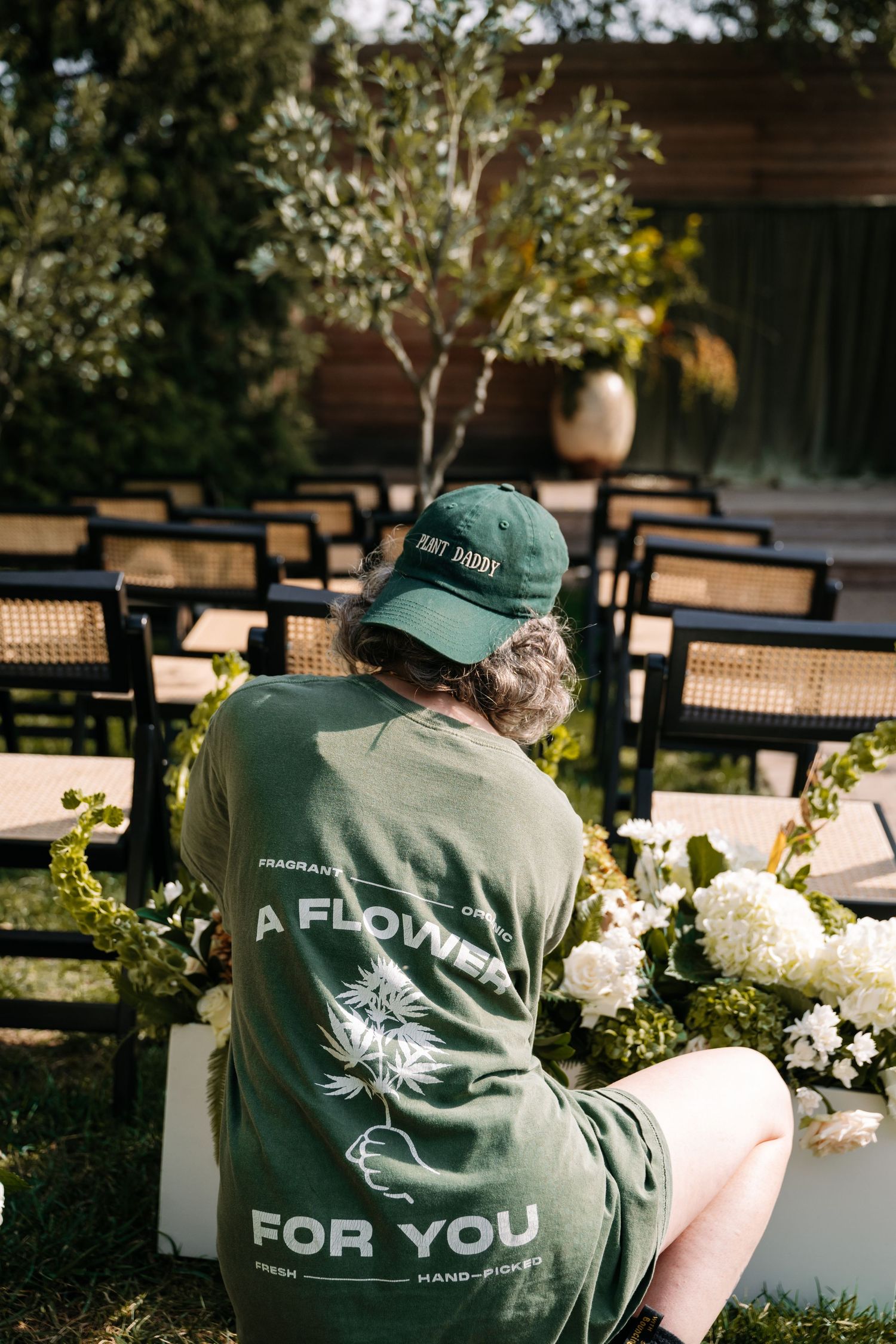 Person wearing green sweatshirt with text sits on outdoor patio bench near white flowers.