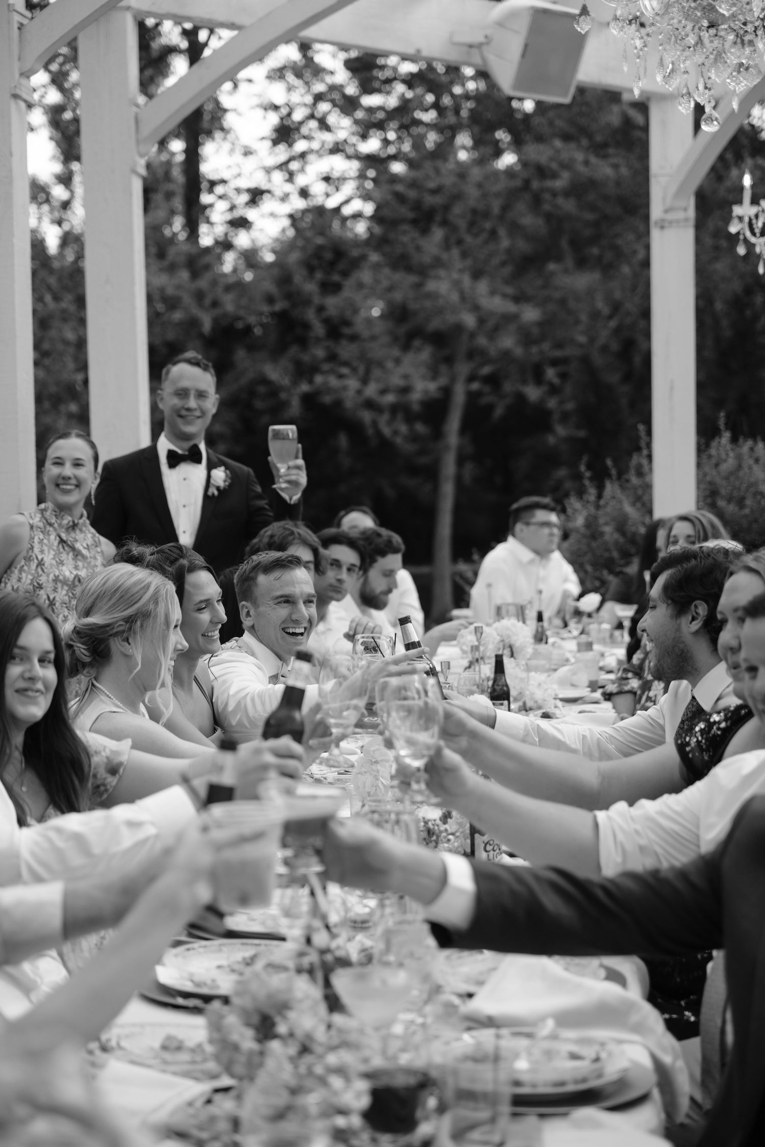 Wedding guests raise their glasses in a toast during an outdoor reception under a white pergola.
