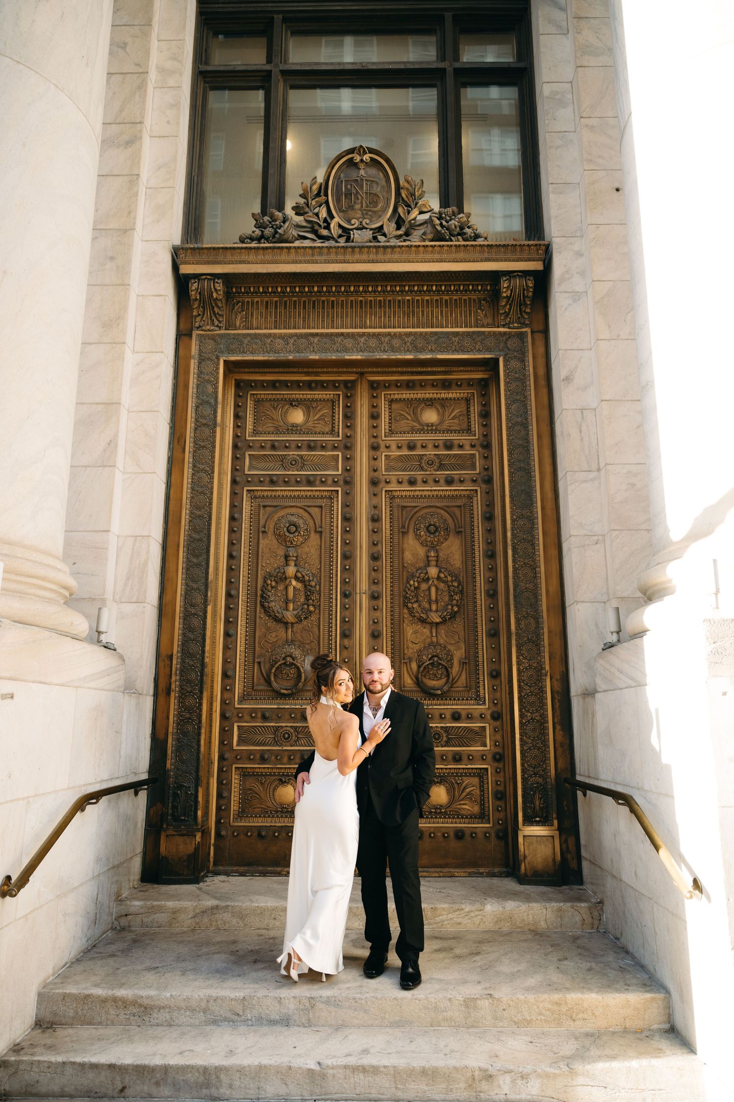Bride and groom embracing on steps before ornate bronze doors of elegant historic building.