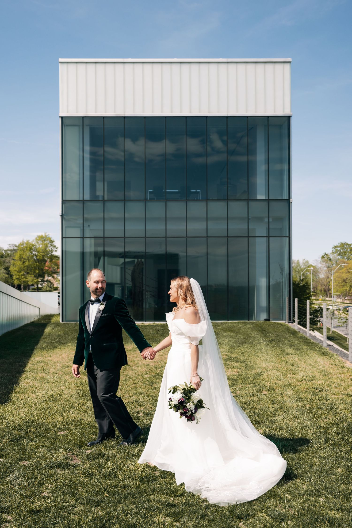 Wedding couple poses against modern glass building exterior with green lawn.