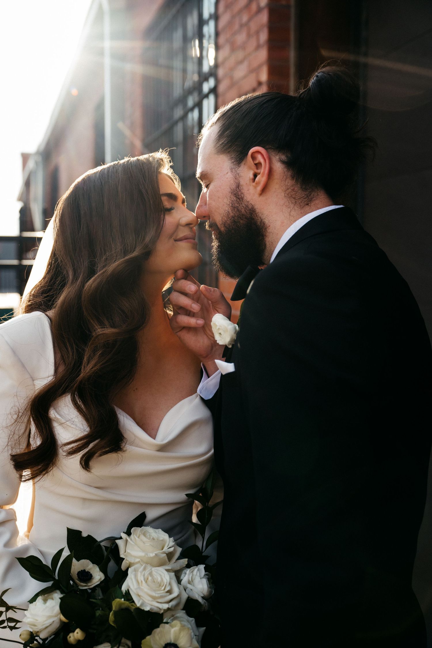 Romantic sunlit moment between couple with white roses bouquet against brick wall.