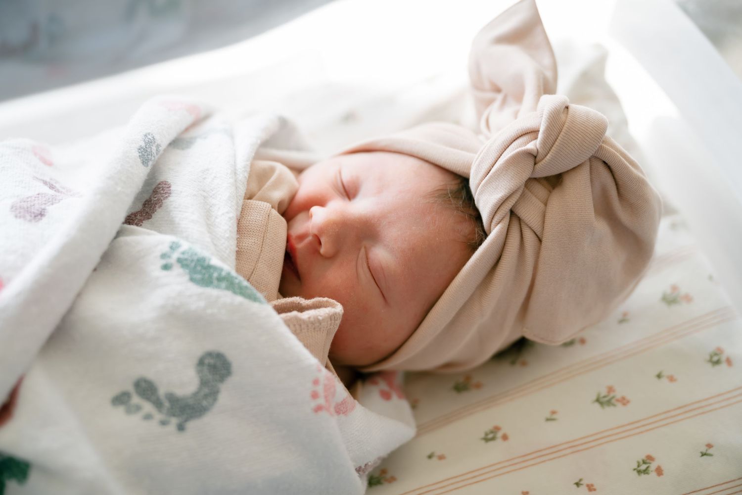 Newborn baby sleeping peacefully wrapped in a floral blanket wearing a beige bow headband in a hospital bassinet.