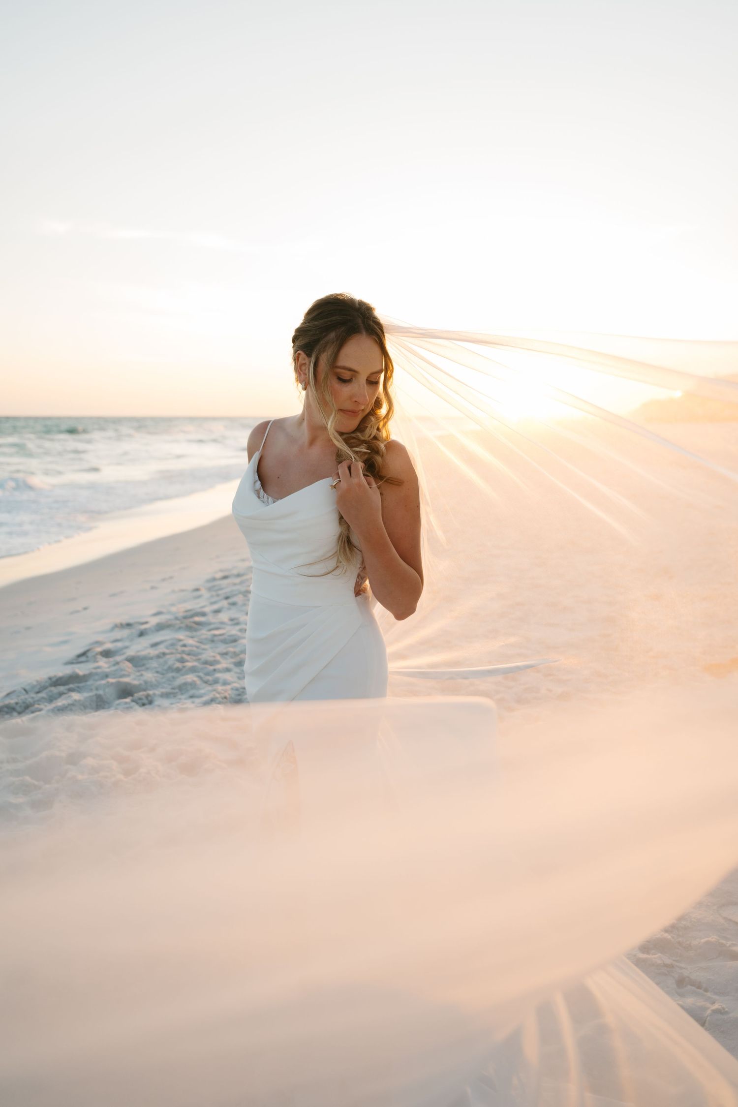 Bride in white gown and flowing veil on a sandy beach at golden sunset with ocean waves behind her.