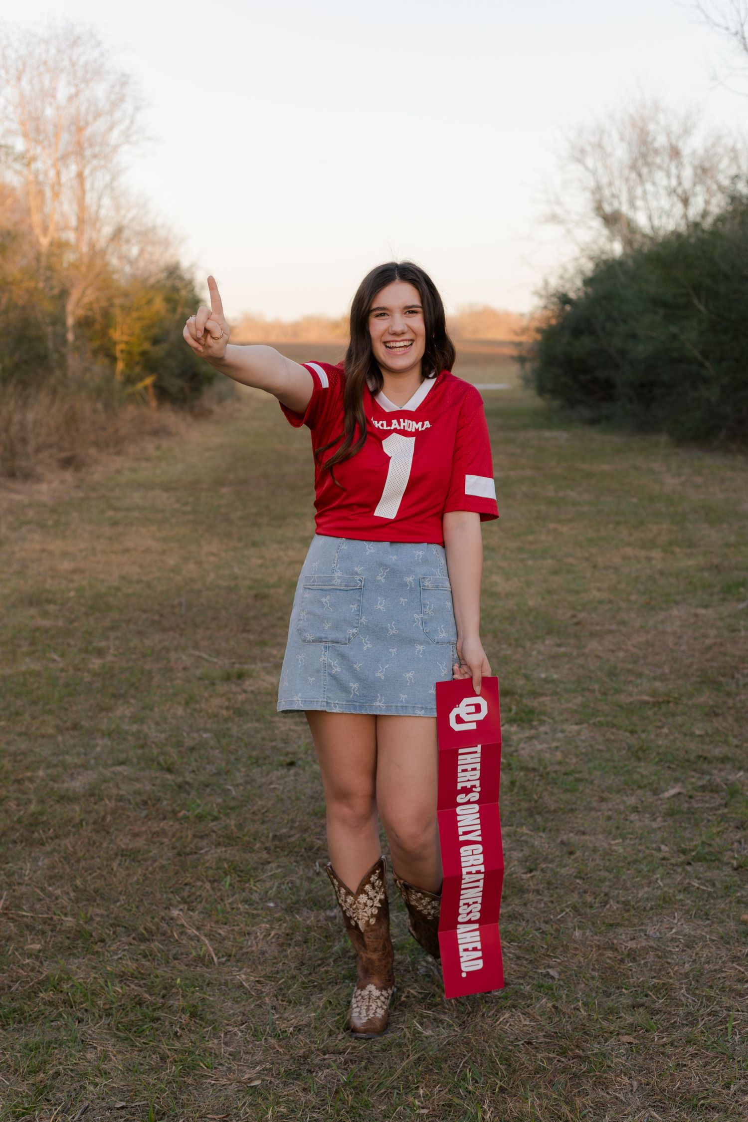 high school senior girl smiles and holds up one finger while holding OU banner and wearing an oklahoma university jersey in a park