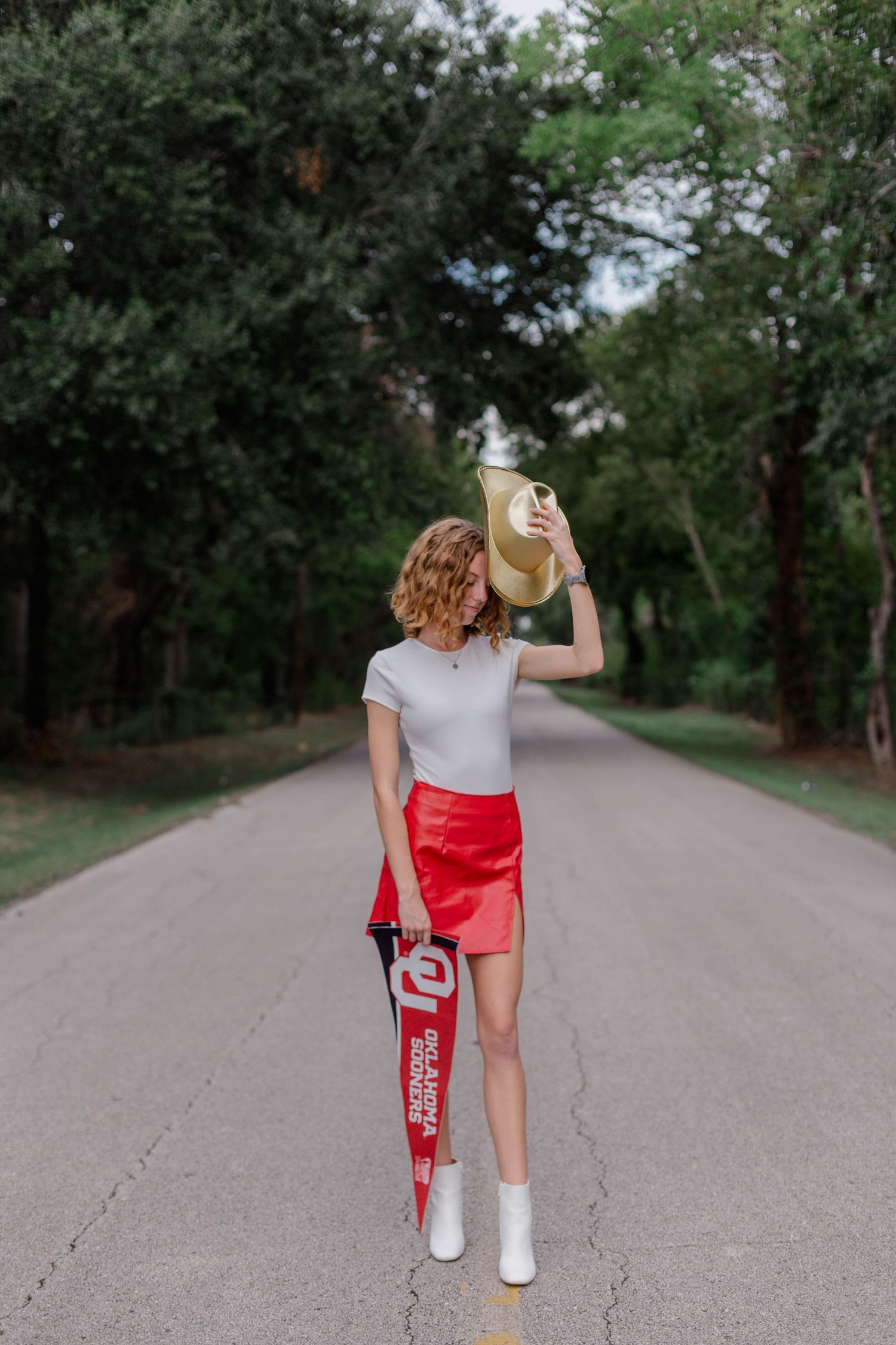 red headed high school senior girl poses with Oklahoma university banner and gold cowboy hat for senior pictures photoshoot