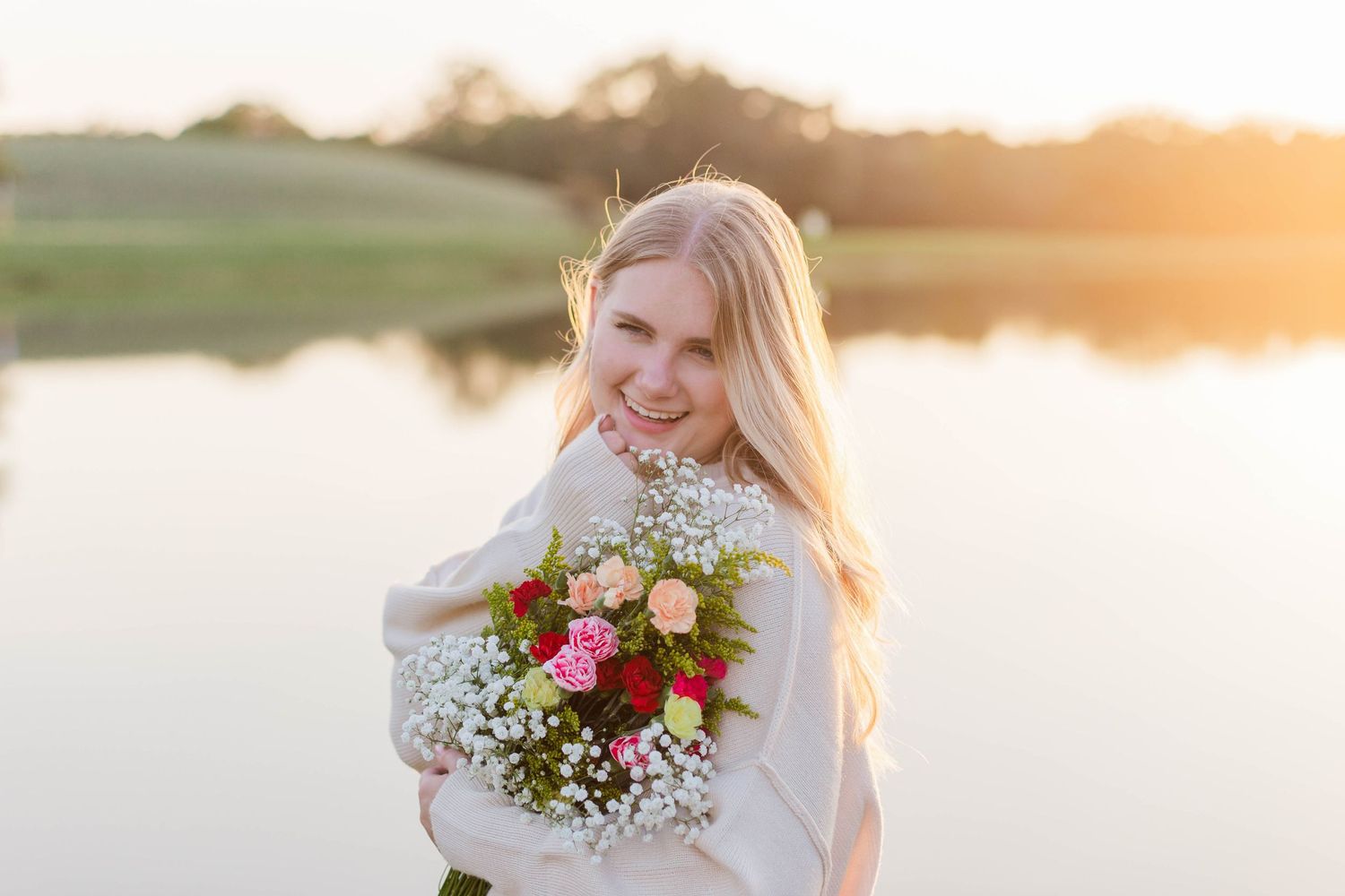 blonde white high school senior girl poses and smiles with a bouquet of roses in front of a pond at sunset in a park in friendswood Texas