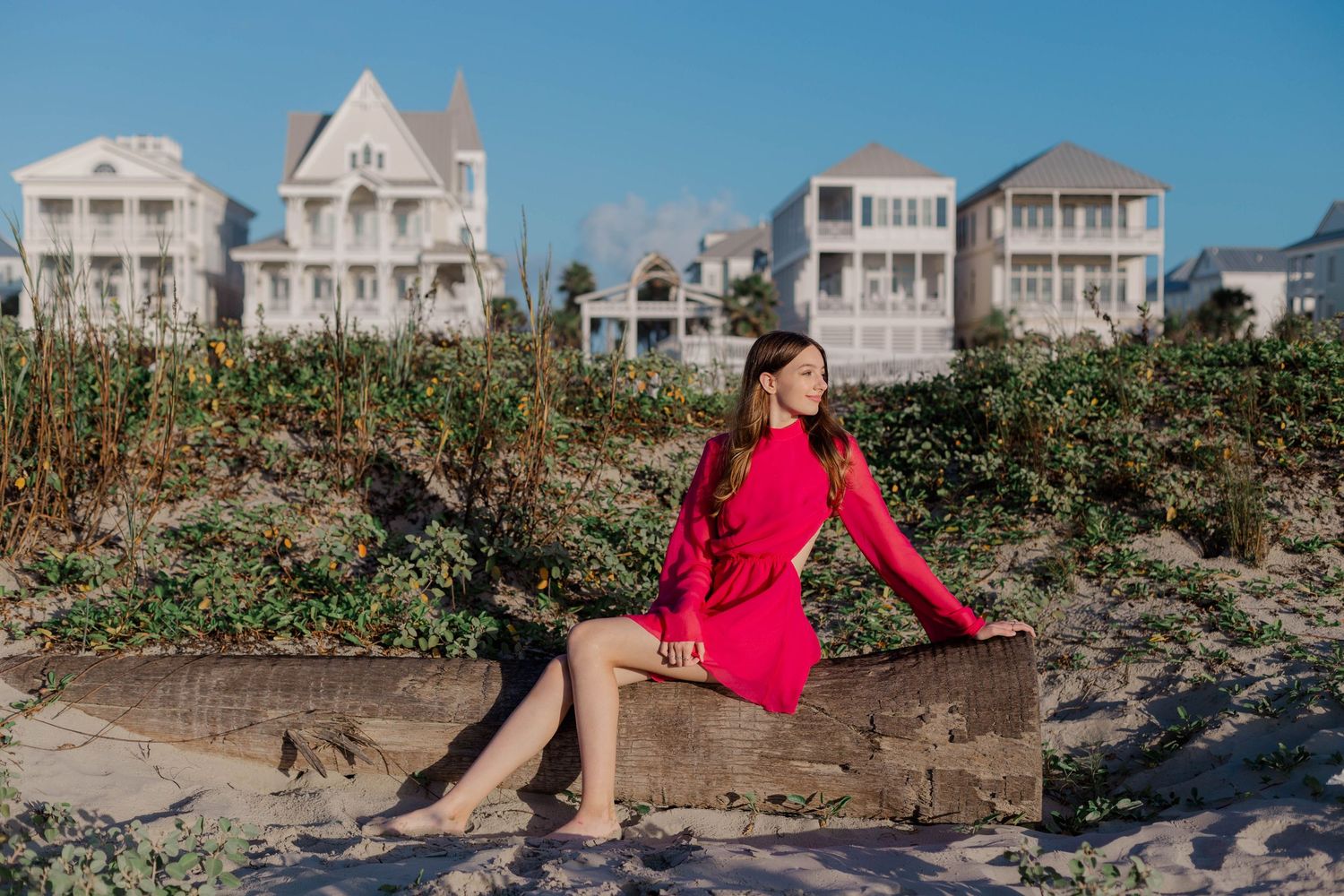 young girl poses for her sweet sixteen photos in front of beach houses on a driftwood log in Galveston Texas