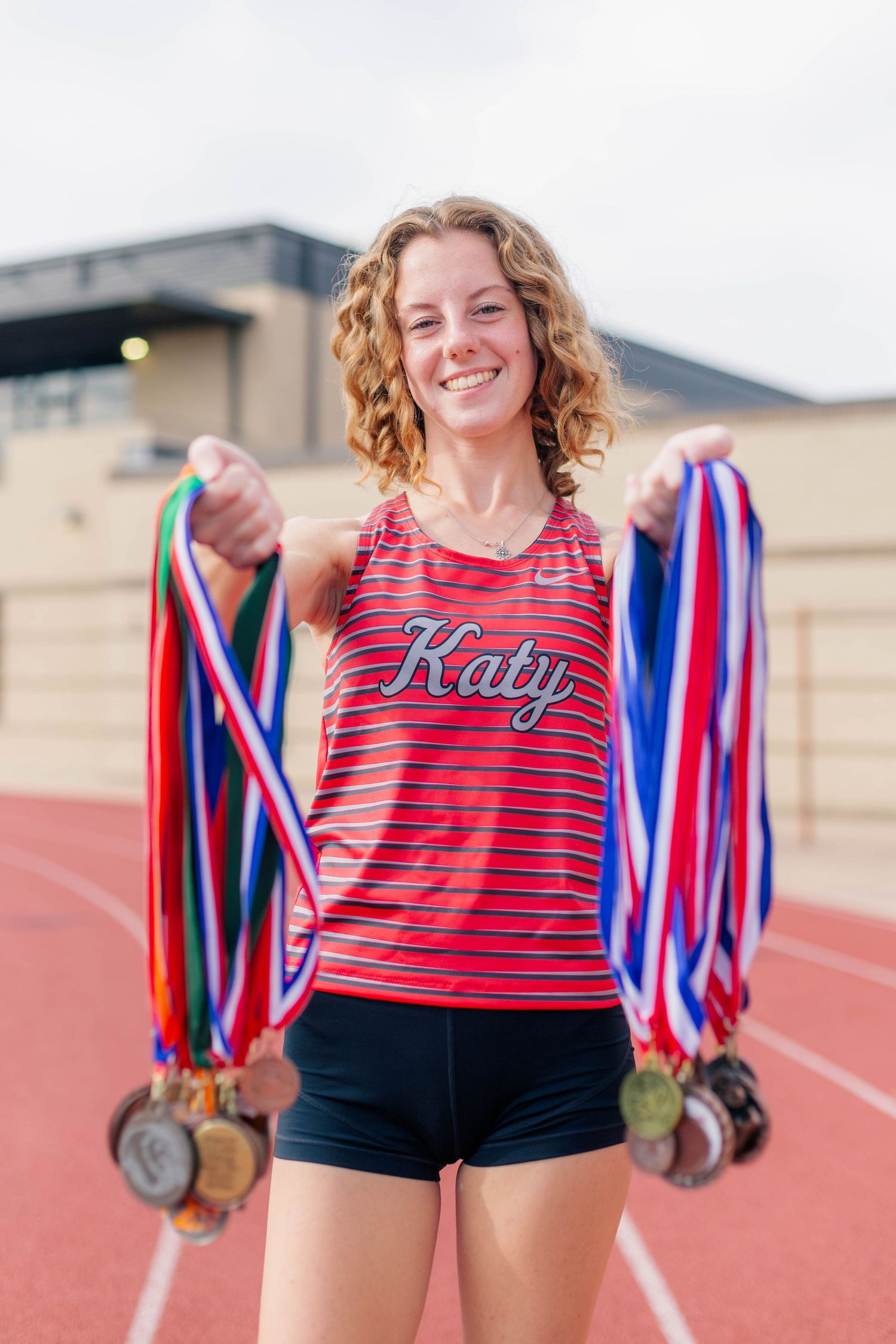 red headed high school senior girl poses with Katy high school track medals on a high school track for senior photos