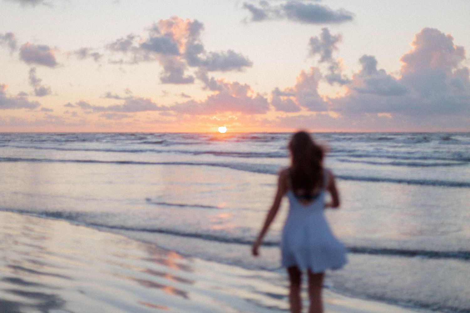 girl in white dress walks toward the water at the beach at sunrise in east beach in Galveston Texas for high school senior photos