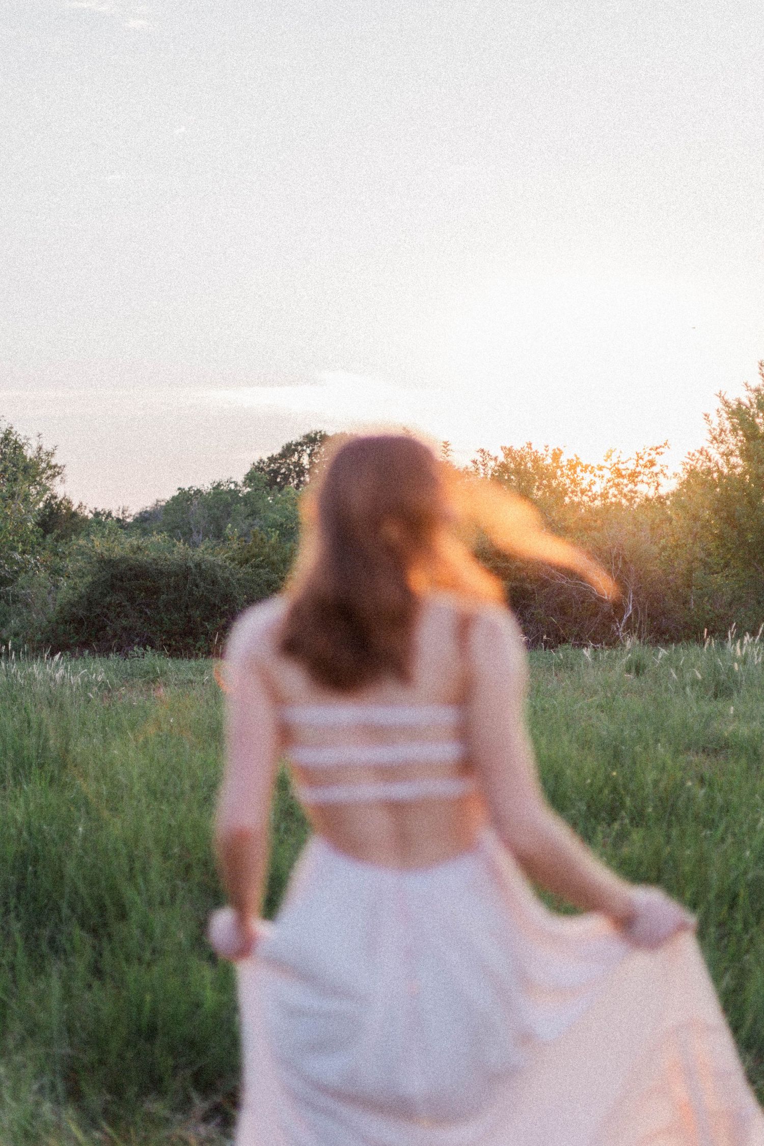 High school senior girl in field of green grass and trees at golden hour sunset in pink dress with red hair flowing in the breeze