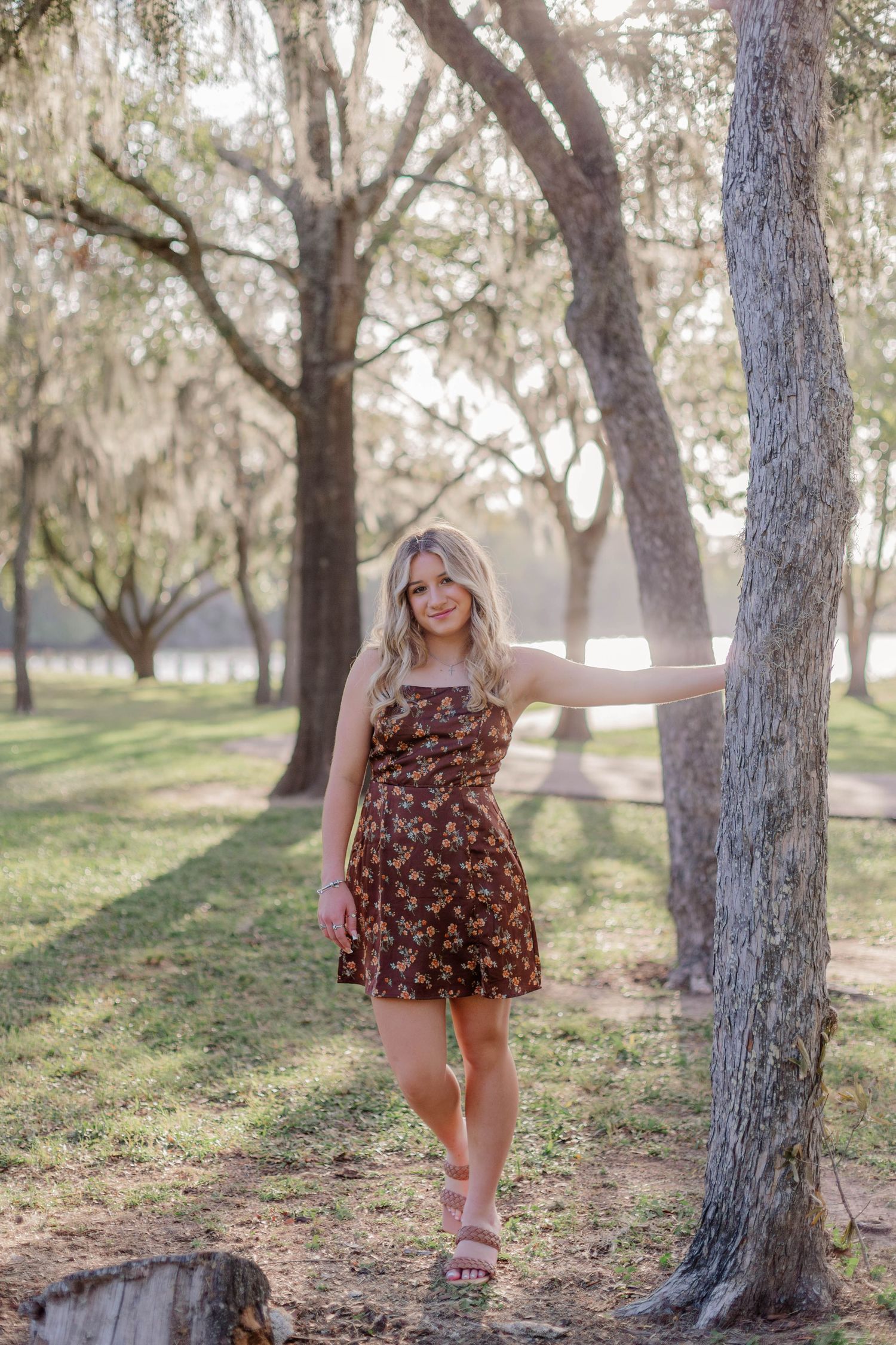 high school senior girl at a park in friendswood Texas holding onto a tree trunk with the sun setting behind her as she smiles