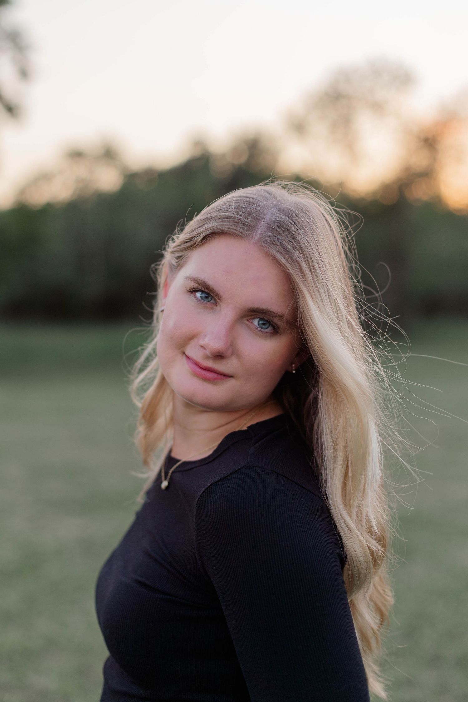blonde high school senior smiles for portrait at sunset in a park in friendswood Texas in a black long sleeve top