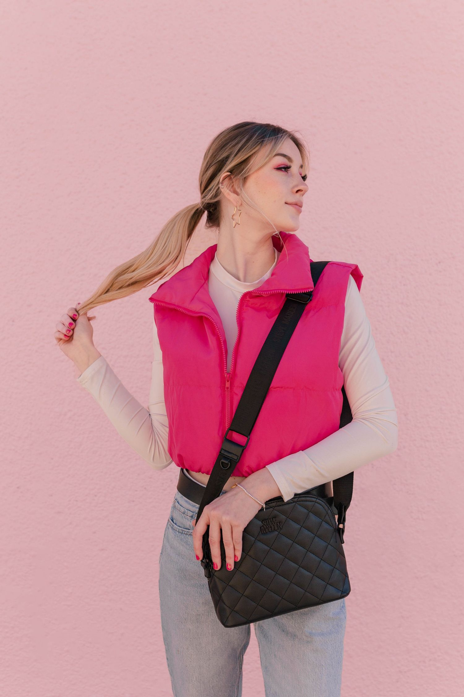 blonde model in hot pink vest with black purse posing as barbie in front of a pink wall for a barbie themed photoshoot