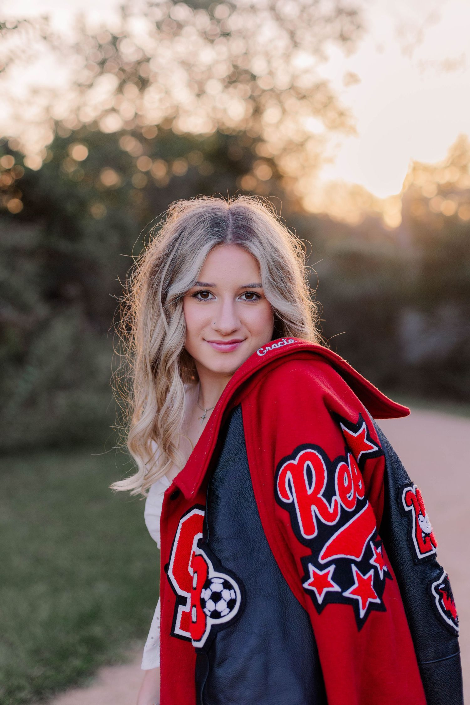 high school senior girl poses with letterman jacket draped over her shoulder at sunset in a park in friendswood Texas for senior photos