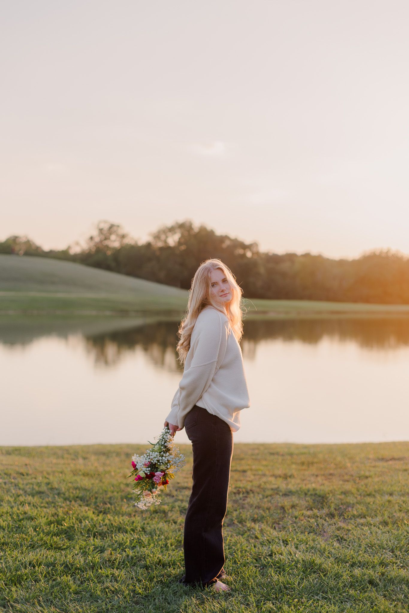 white blonde high school senior girl holds bouquet behind her back in stands in front of pond at sunset for senior photos in friendswood