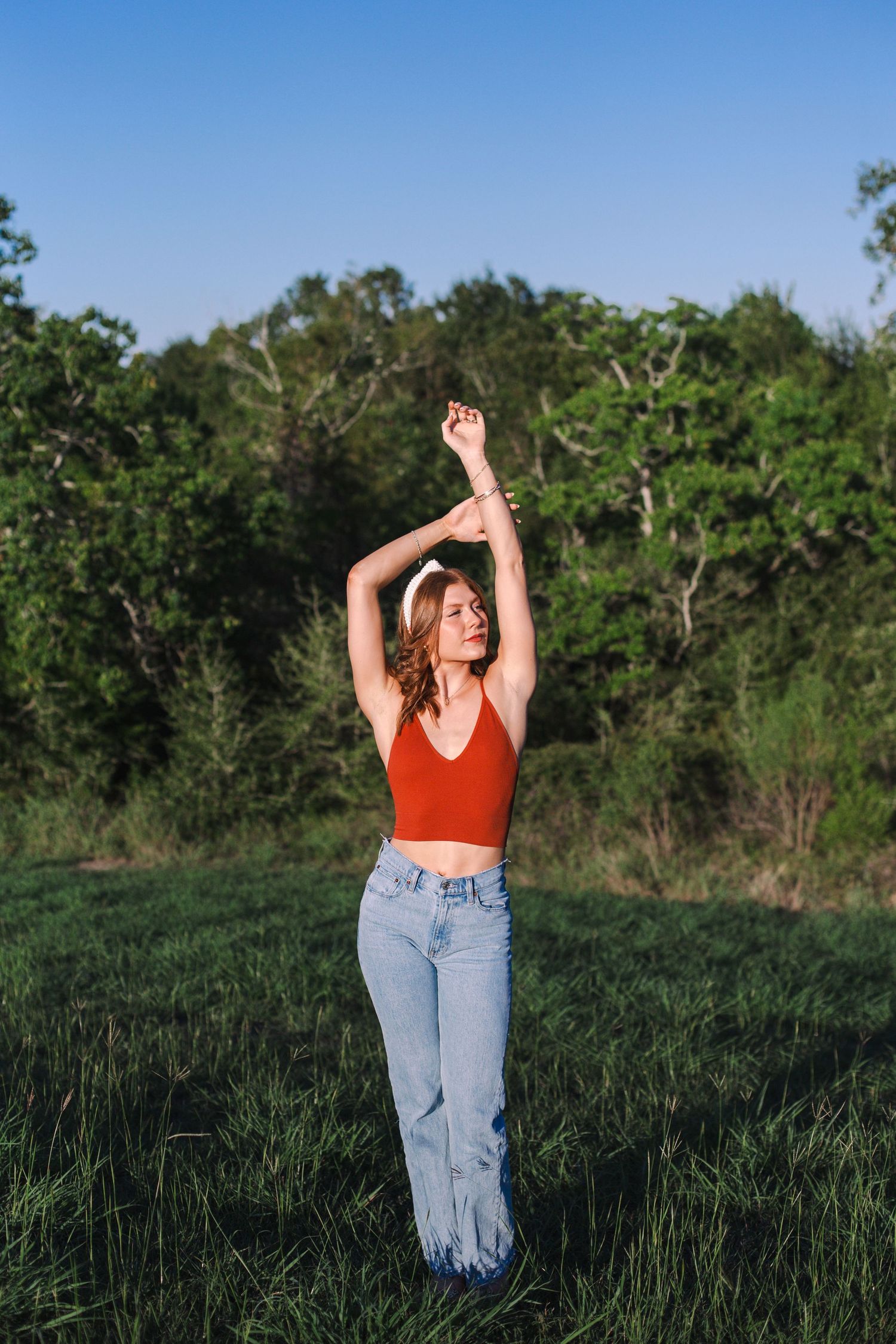 girl poses for senior photos in a field of green grass with green trees behind her