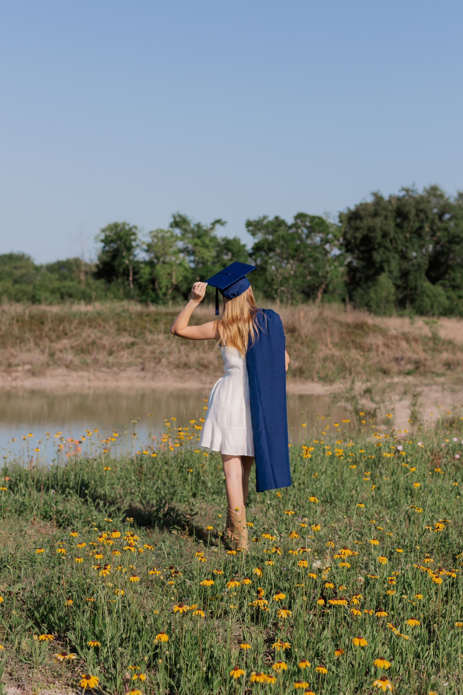 blonde white high school senior girl poses with cap and gown in field of wildflowers for senior pictures photoshoot in Alvin Texas