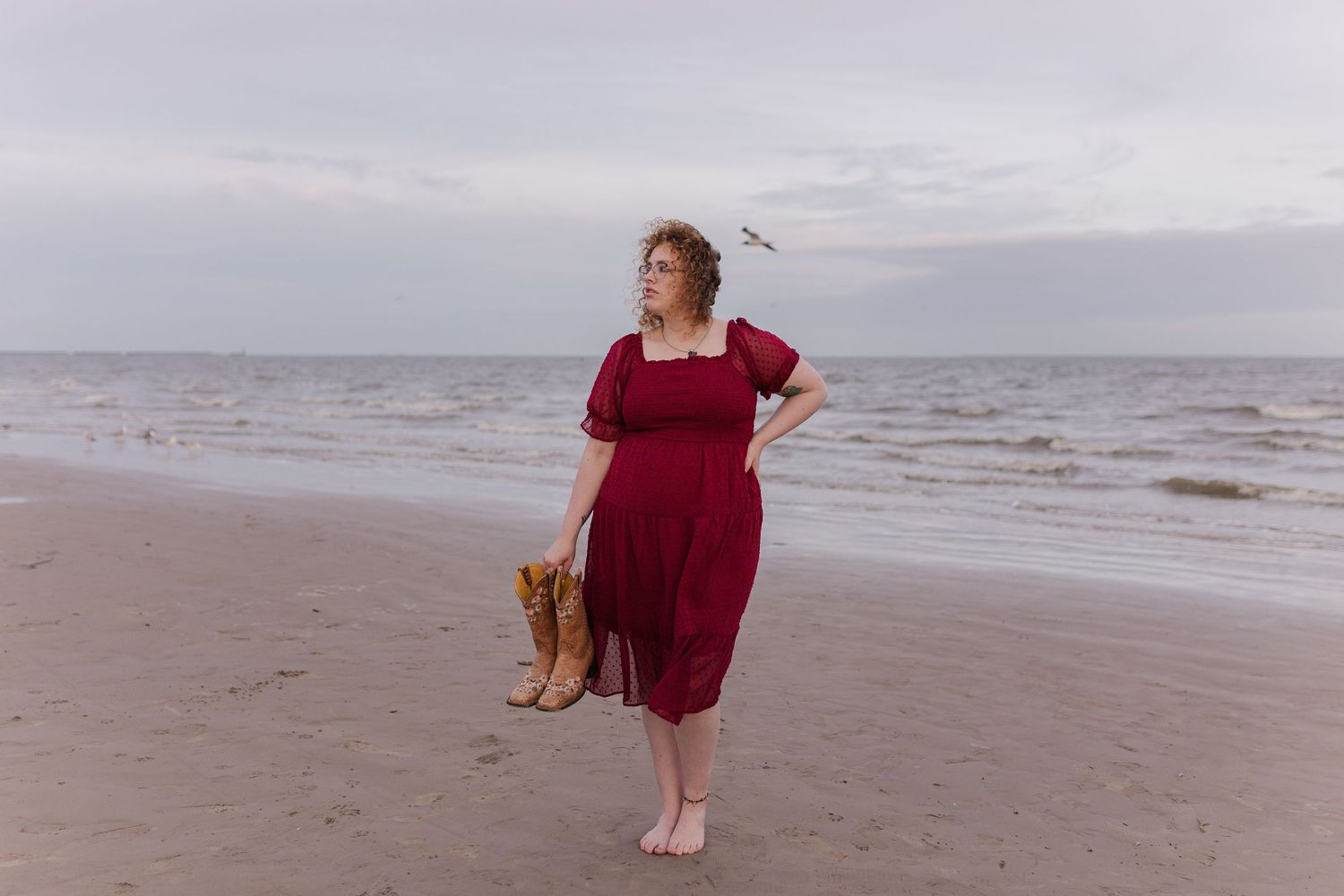 high school senior girl poses on beach while holding cowboy boots in seabrook Texas
