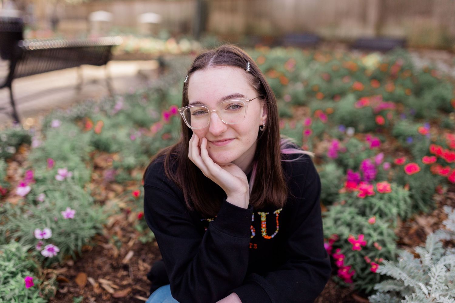 white brunette high school senior girl smiles at camera in front of a small flower bed in a black sweater for senior pictures photoshoot