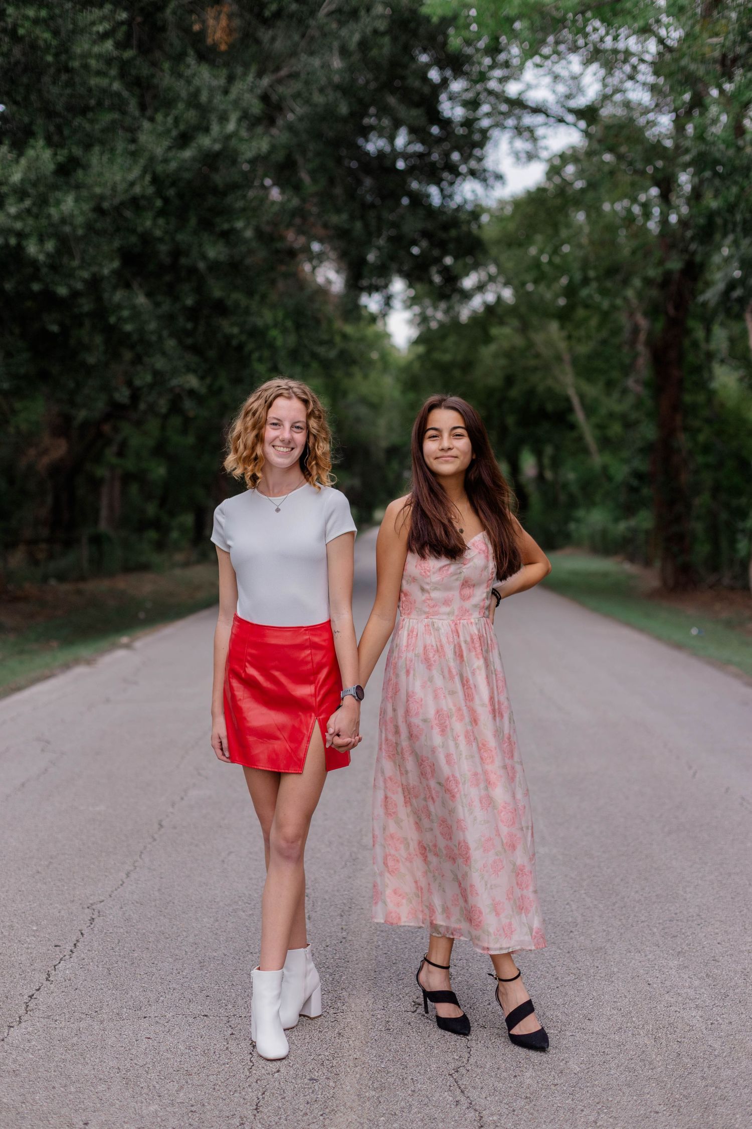 white girl and hispanic girl hold hands and pose for photos on a treelined street for high school senior photos
