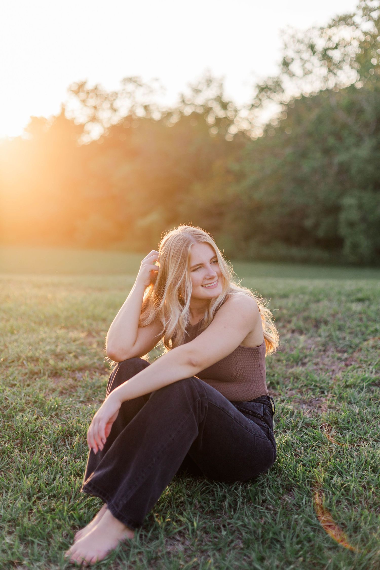 high school senior girl sitting on green grassy hill with trees behind her in a park in friendswood Texas with hand in her blonde hair