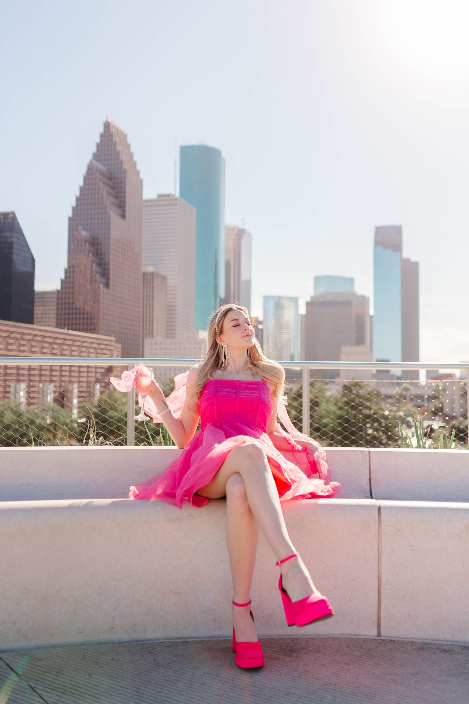 blonde model posing on rooftop of POST Houston in a hot pink dress and heels holding a glass of sparkling juice in barbie themed photoshoot