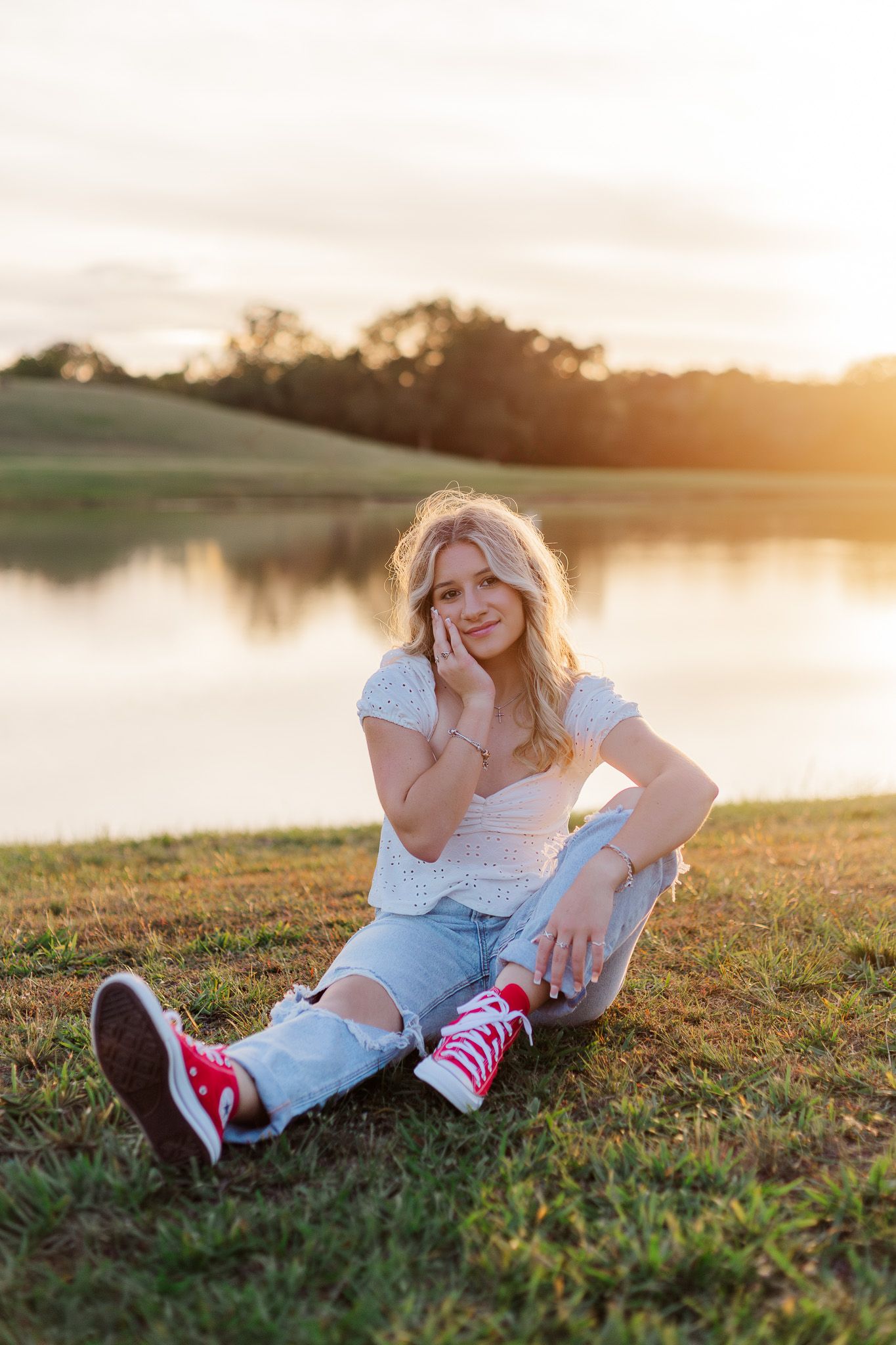white blonde high school senior girl sits in front of pond at sunset for senior photos