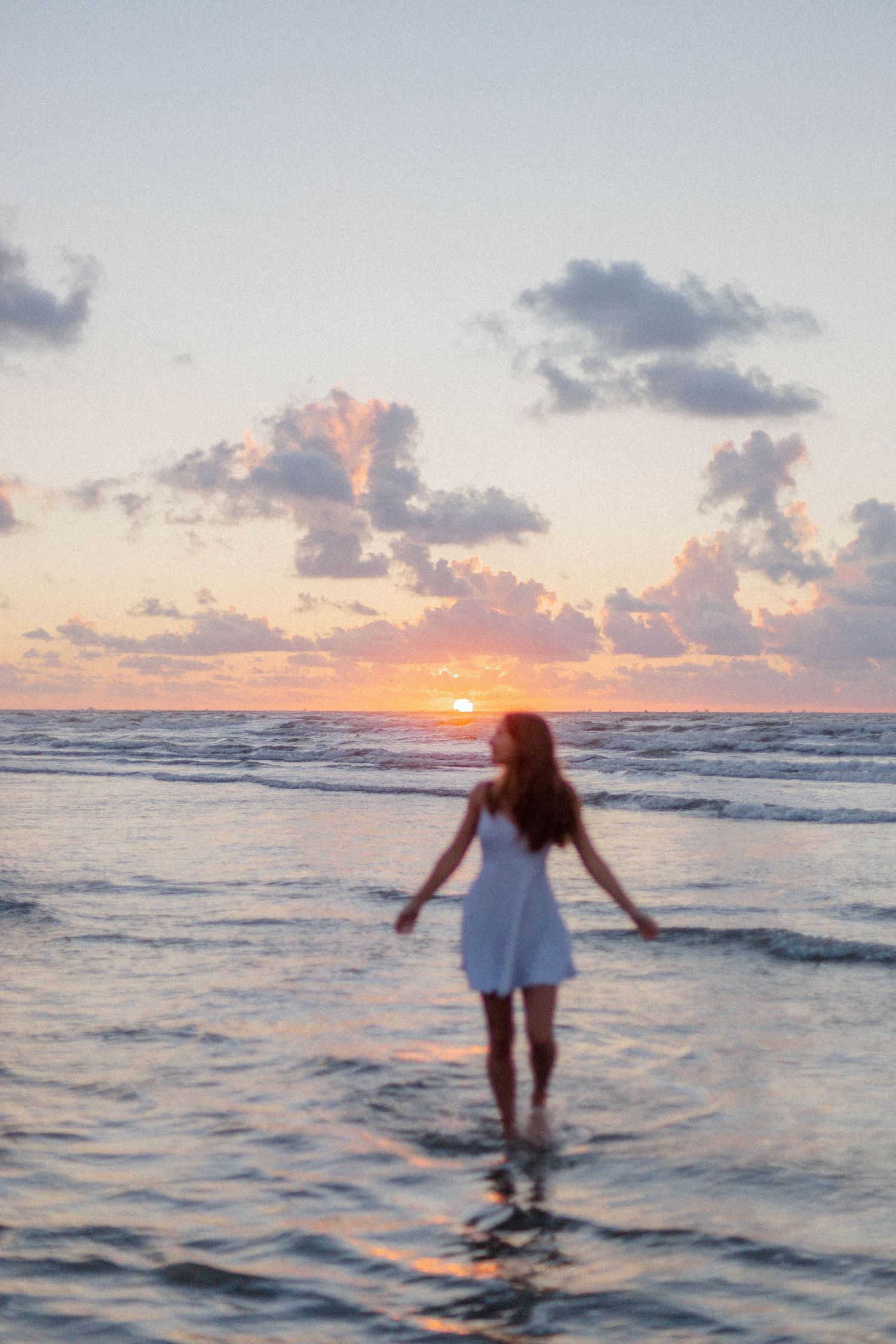 high school senior girl looks up at the sky during sunrise at the beach in the water for senior photos