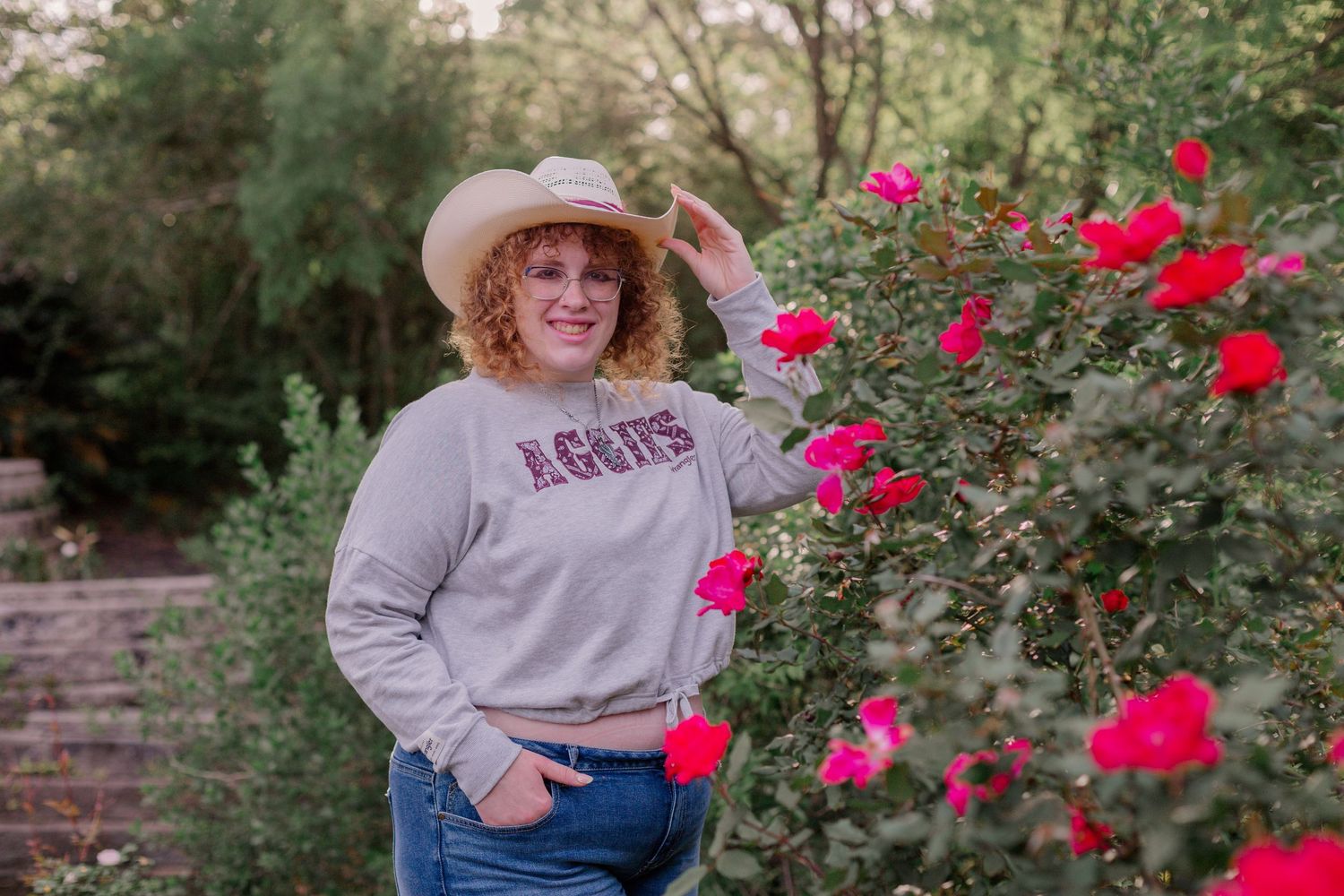 high school senior girl wears cowboy hat and wears an Aggie sweater for senior photos next to a hot pink rose bush