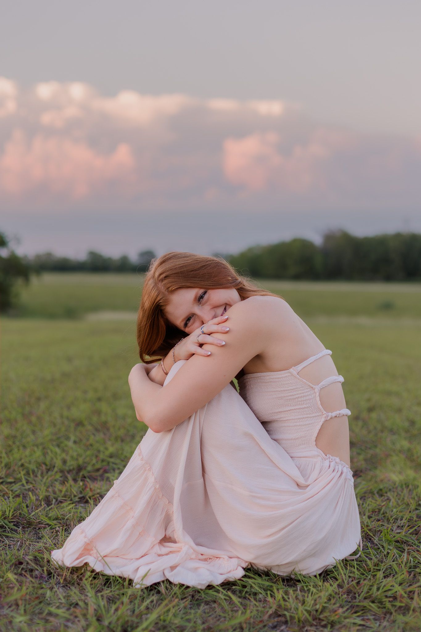 white red head high school senior girl sits in the green grass, hugging her knees and smiling in front of pink clouds at sunset for photos