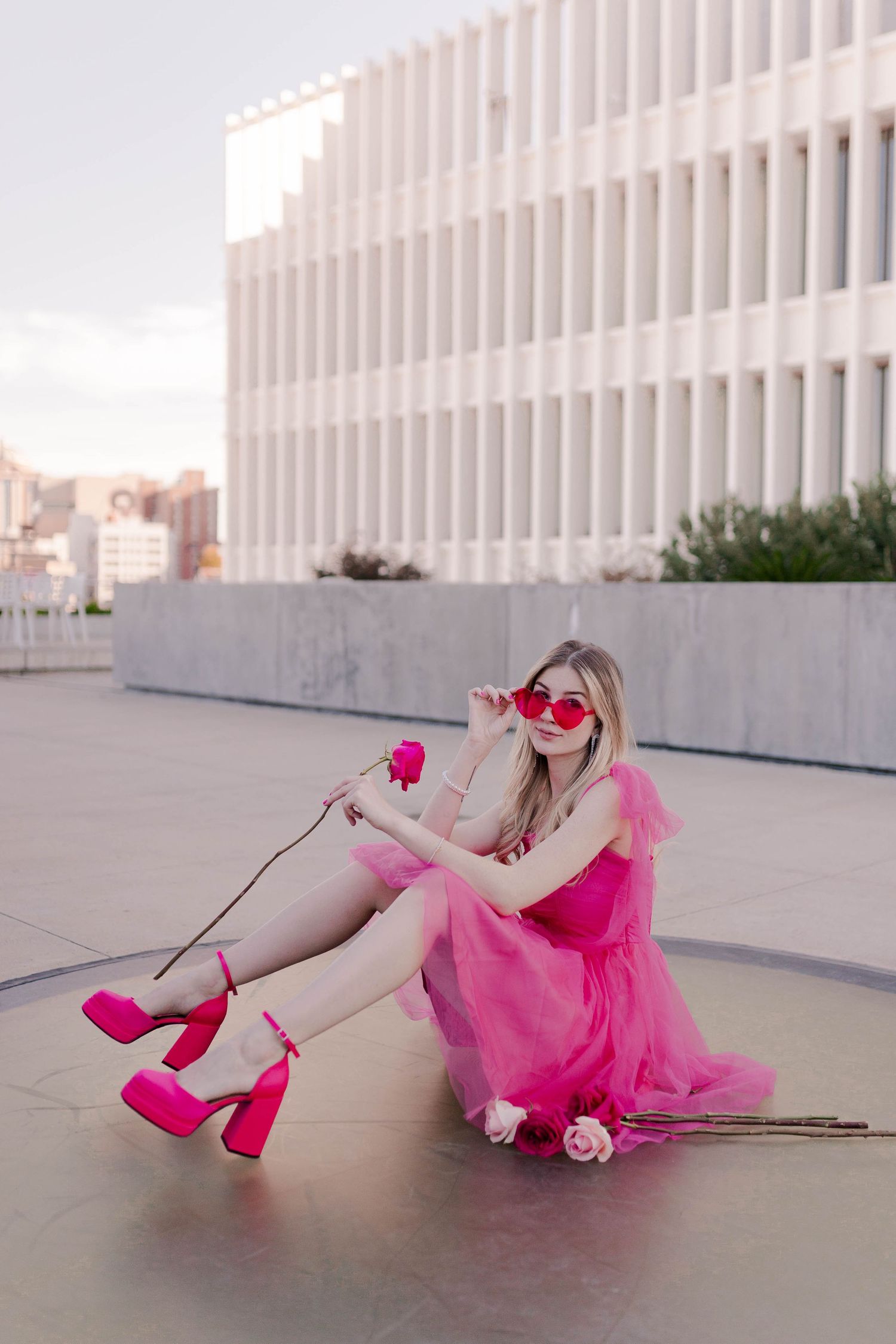 blonde model posing in a hot pink dress with pink heart shaped balloons at POST Houston with red and pink roses at a barbie theme photoshoot