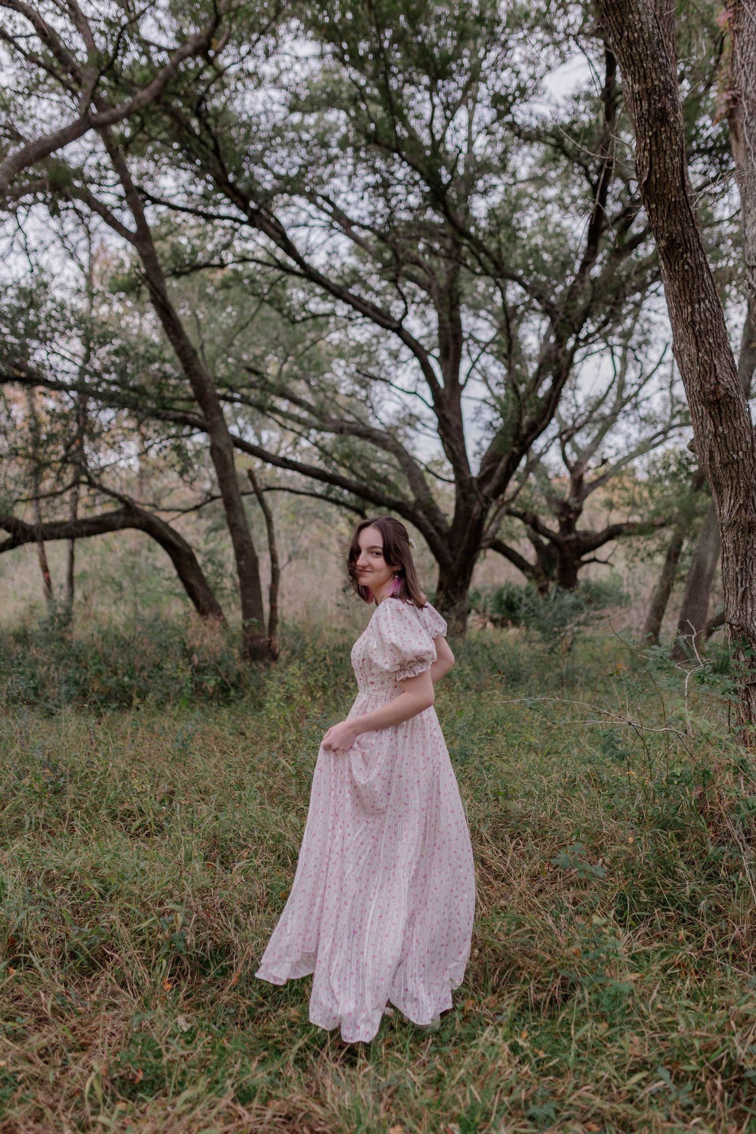 white brunette high school senior girl turns toward camera in a long floral dress and smiles in a forest in friendswood Texas