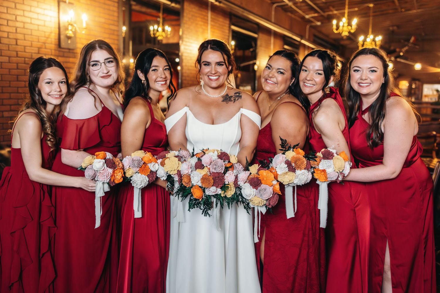 Wedding party in burgundy dresses poses together holding bouquets in front of exposed brick walls.