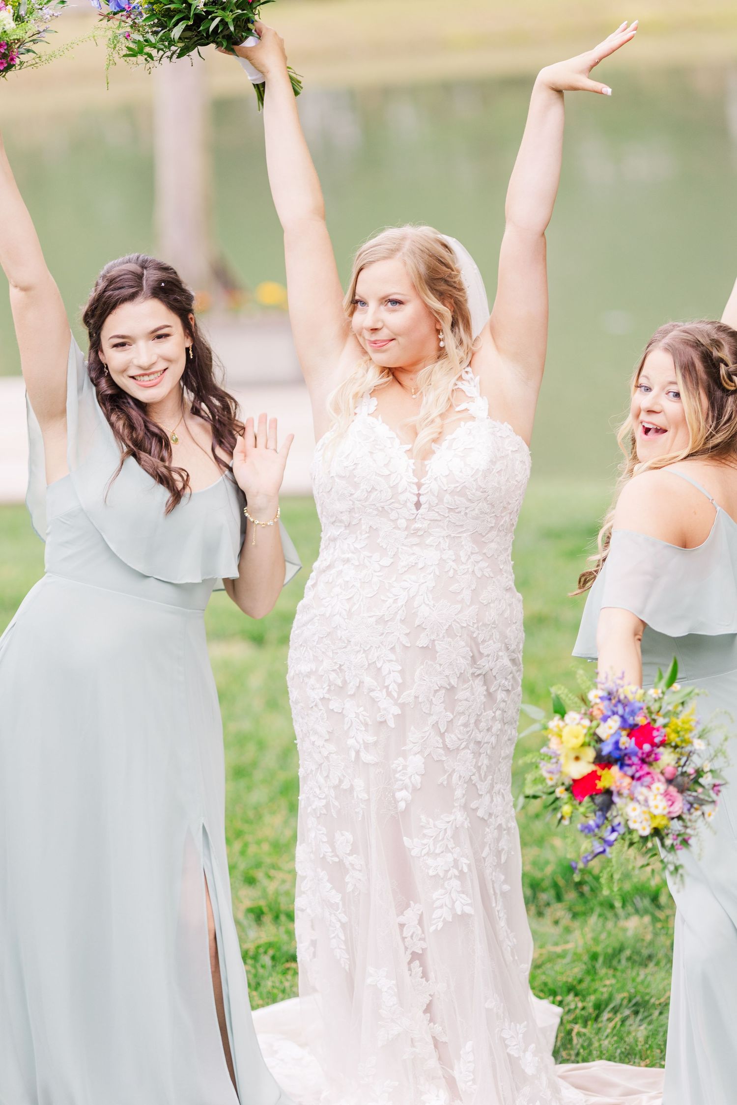 Bridesmaids in light sage dresses celebrate joyfully with the bride in a sunny outdoor setting.