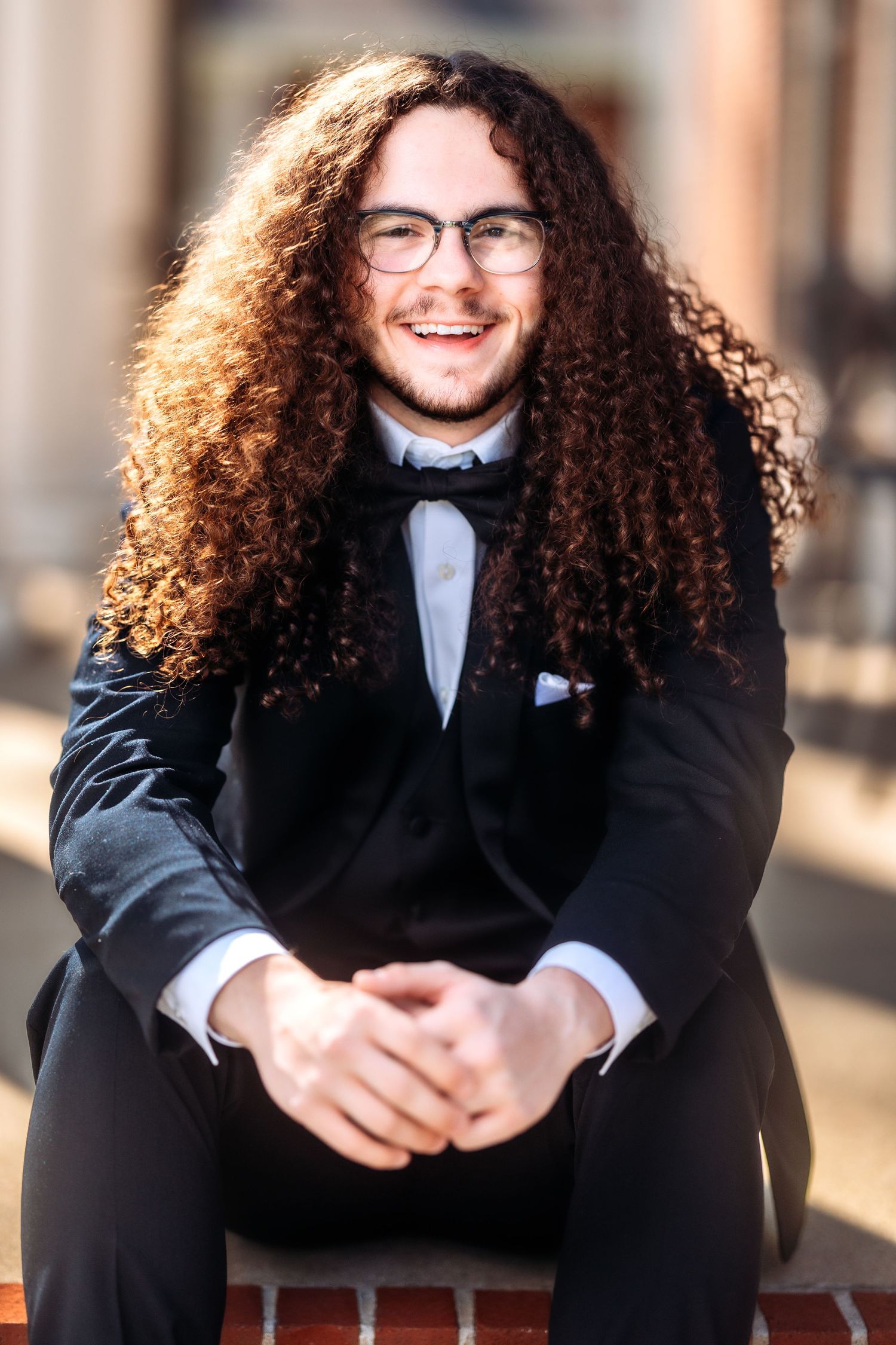 A person with long curly hair wearing glasses, a bow tie and black suit sits smiling on a brick ledge outdoors.