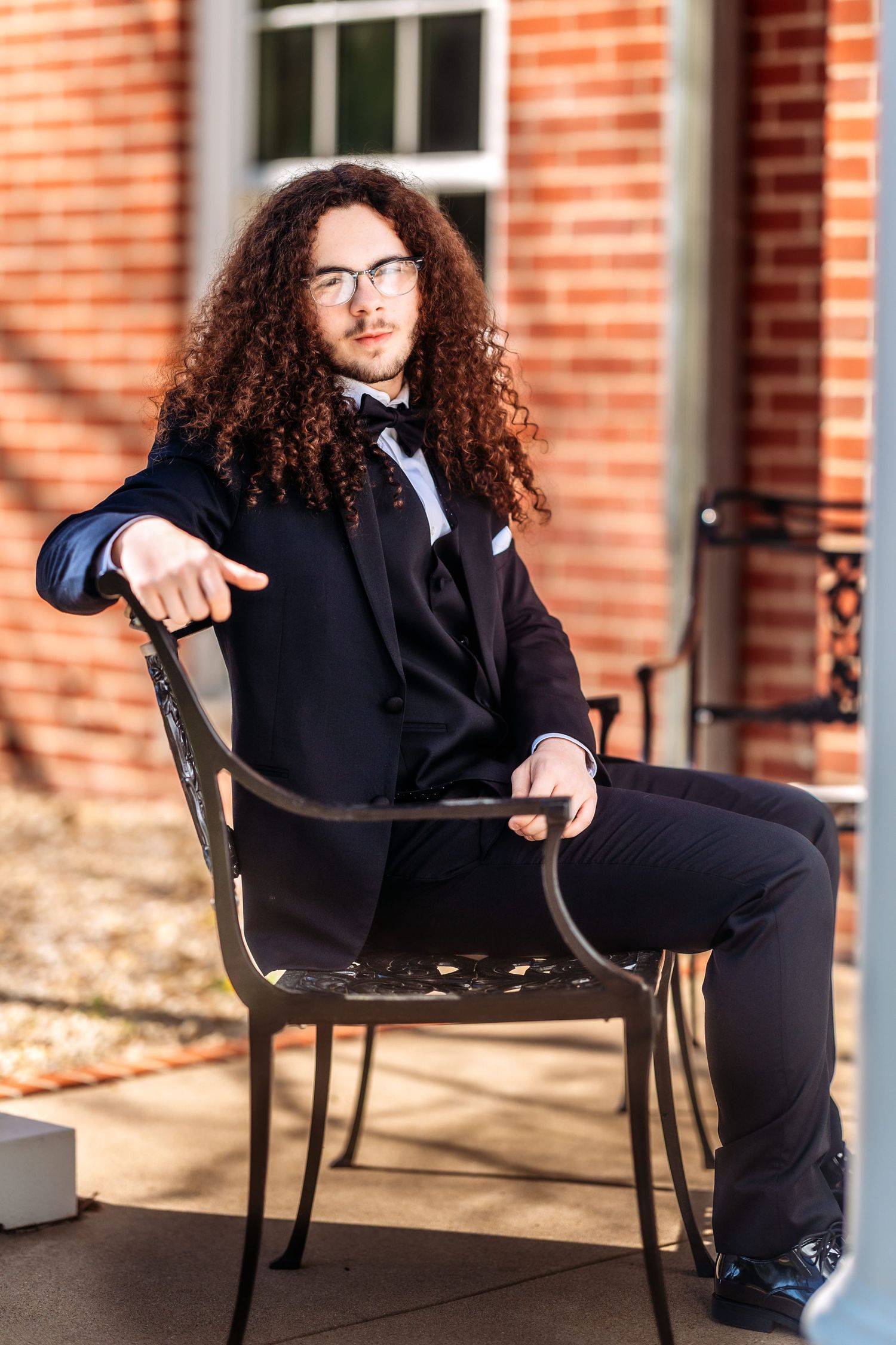 A person with long curly hair wearing a black suit and bowtie sits in an outdoor metal chair against a brick building.