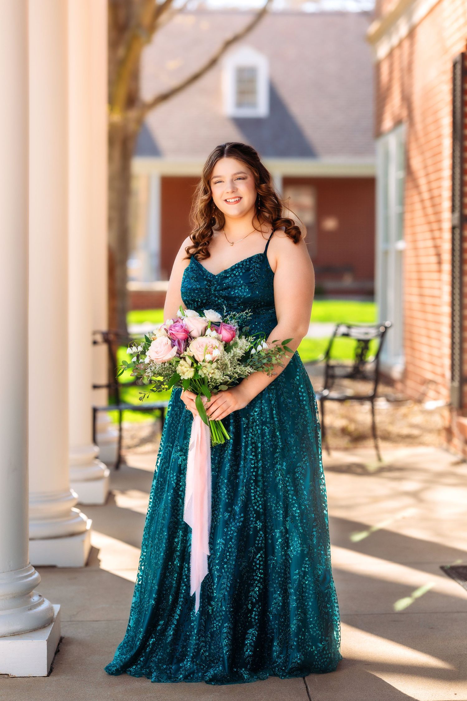 A bridesmaid in an emerald sequin dress poses with a pink and white bouquet on a sunny porch with white columns.