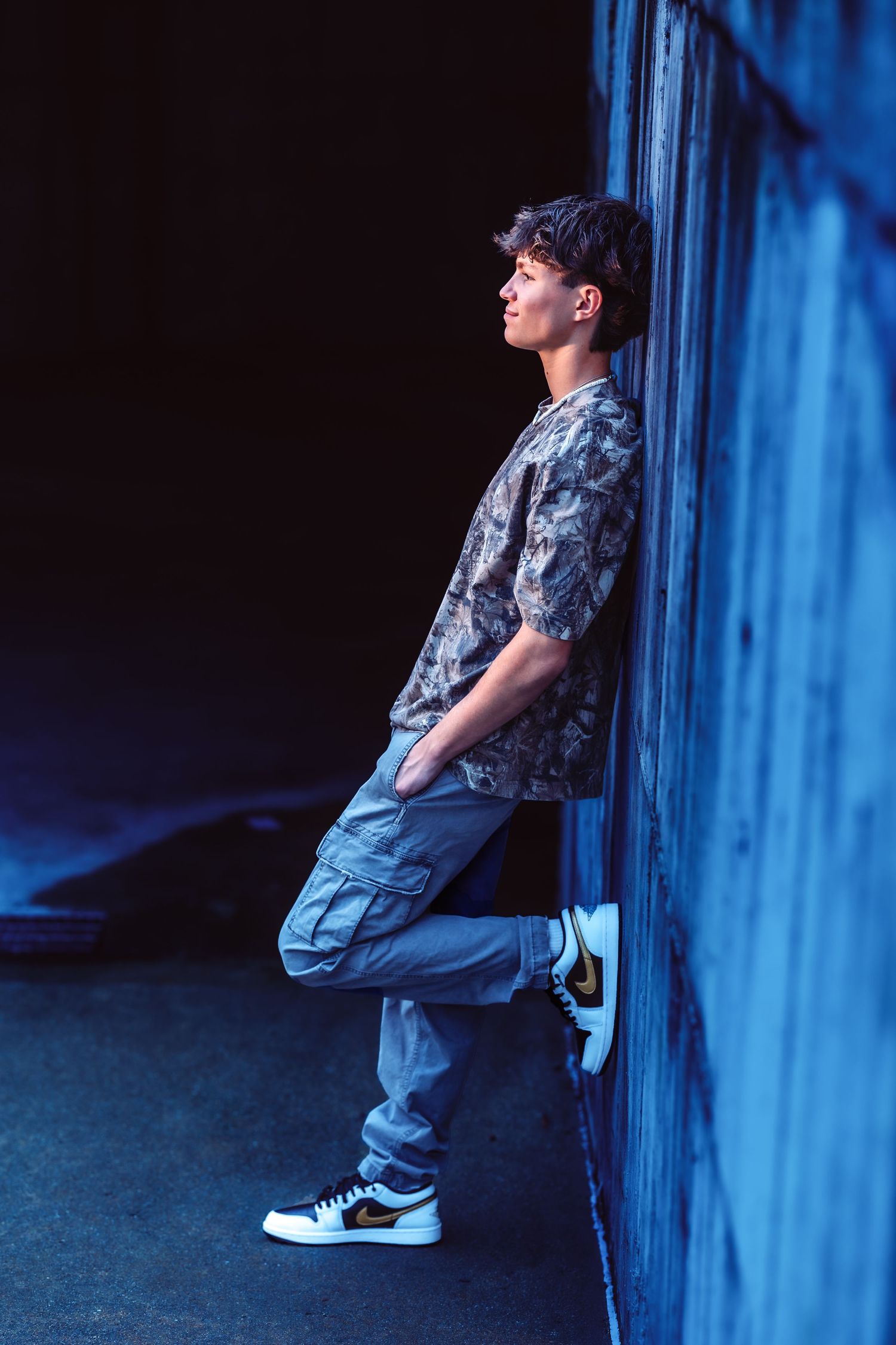 Person in casual wear and sneakers leans against blue wall in moody lighting creating artistic urban portrait series.