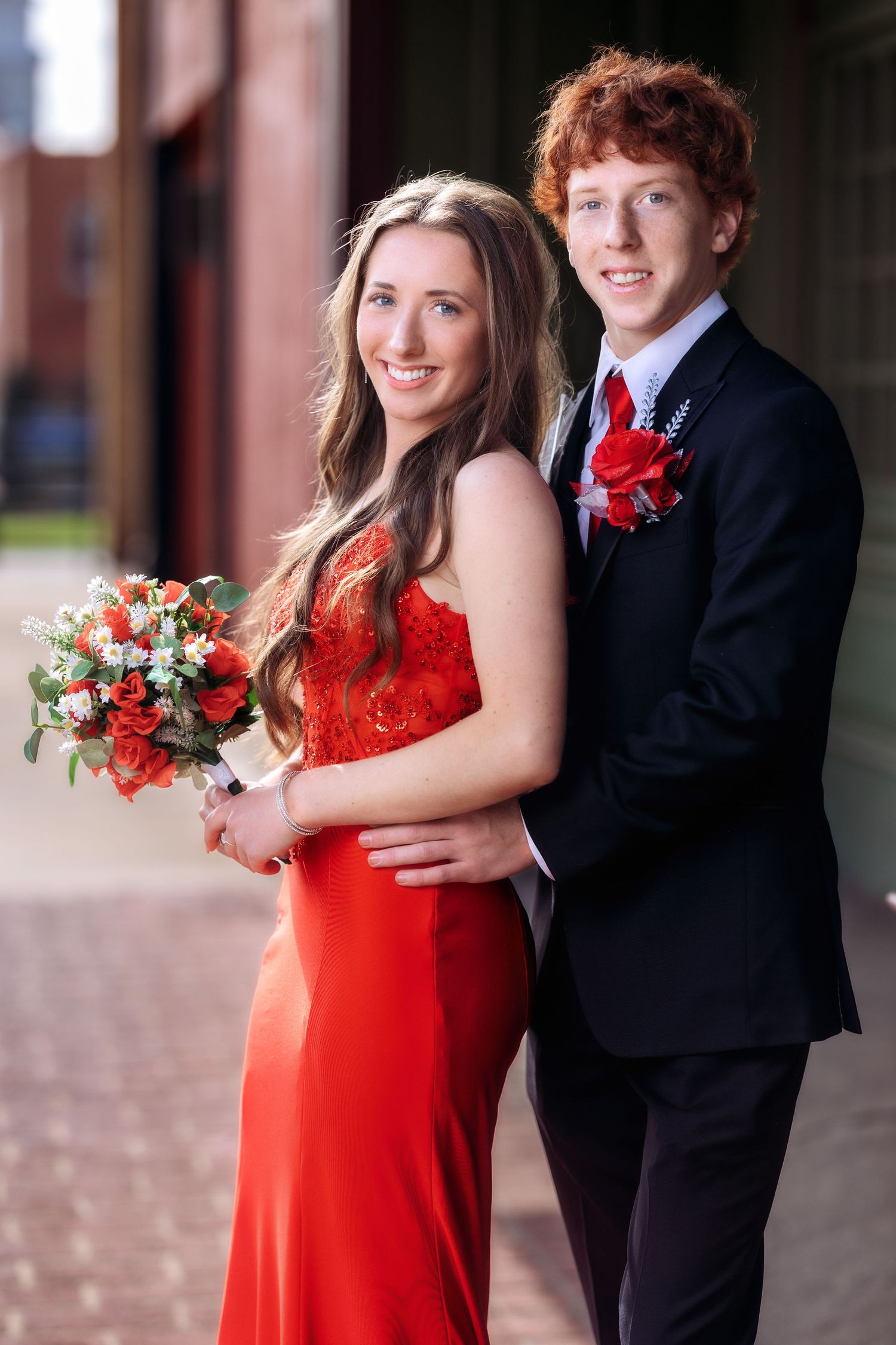 Elegant red dress and black tuxedo formal portraits against brick backdrop.