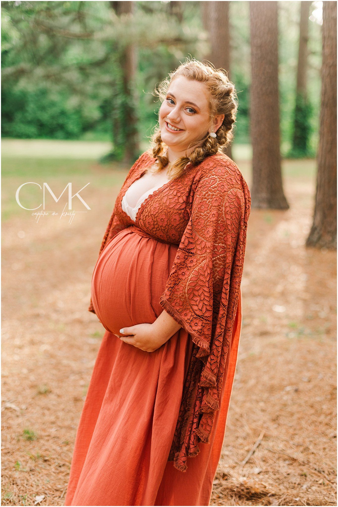 Maternity portrait in a pine forest with a flowing rust-colored dress and lace overlay.
