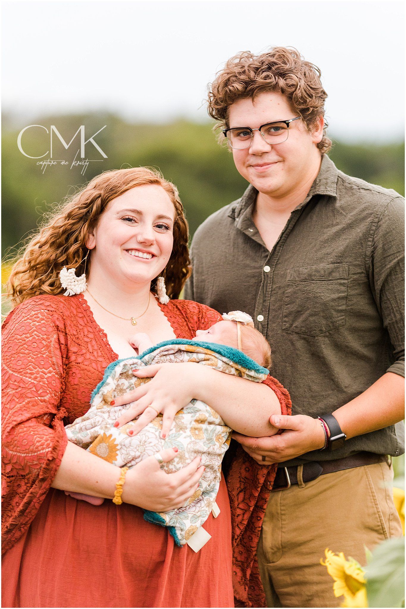 Outdoor family portrait with baby wrapped in patterned blanket against white background.