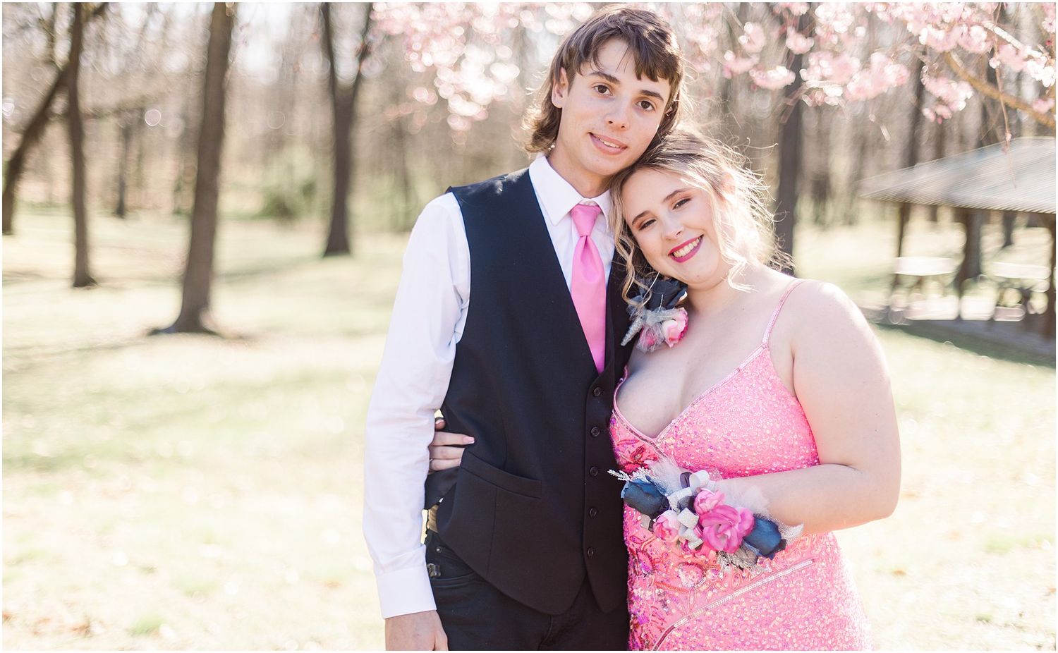 Formal couple poses together under spring cherry blossoms wearing pink and black formal attire.