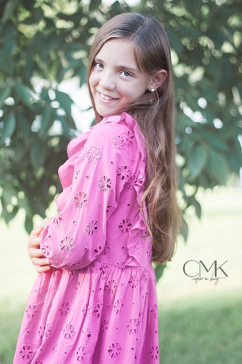 Portrait of young person in pink lace dress posing against leafy outdoor background.