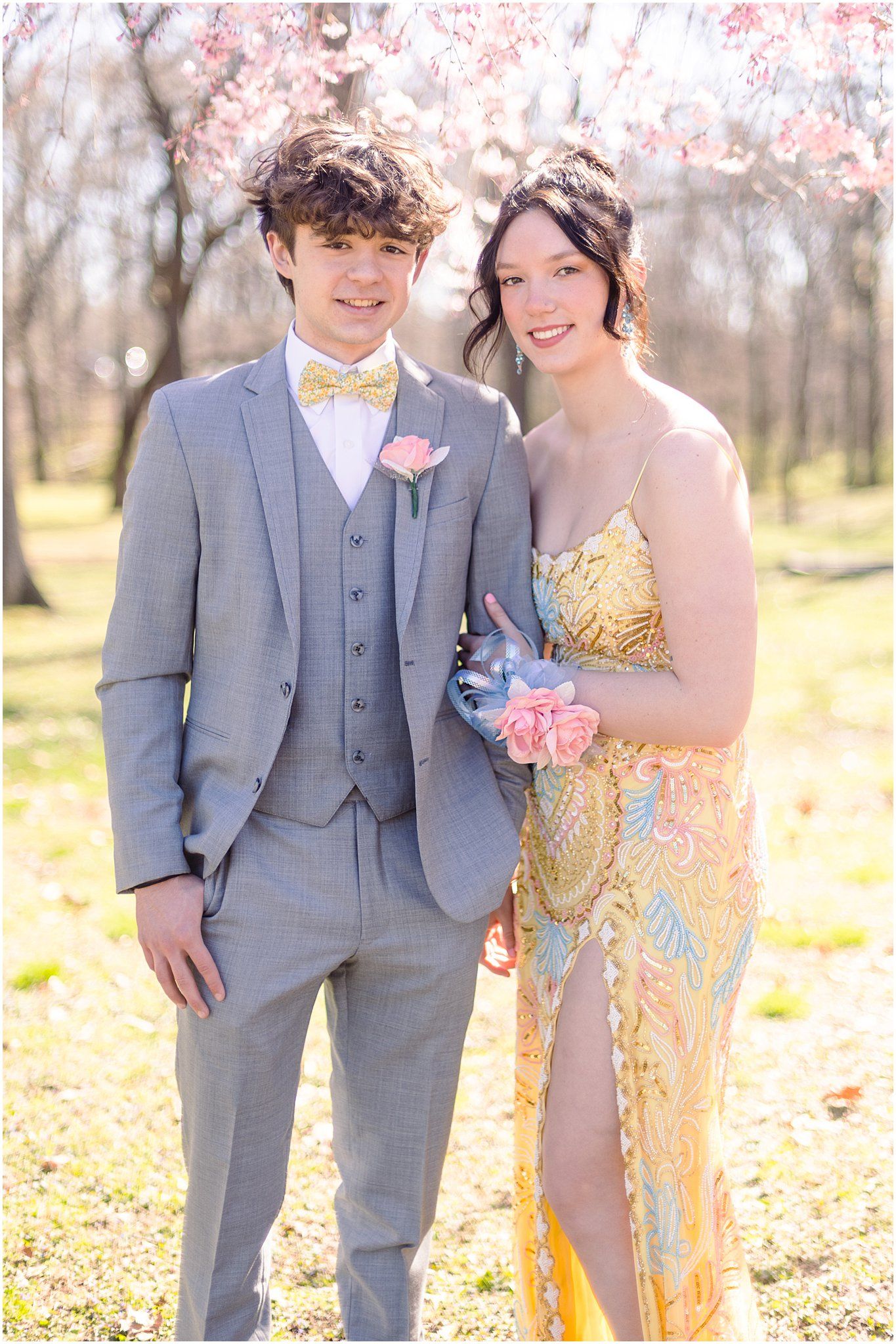 Formal couple pose with pink corsage and boutonniere under blooming trees.